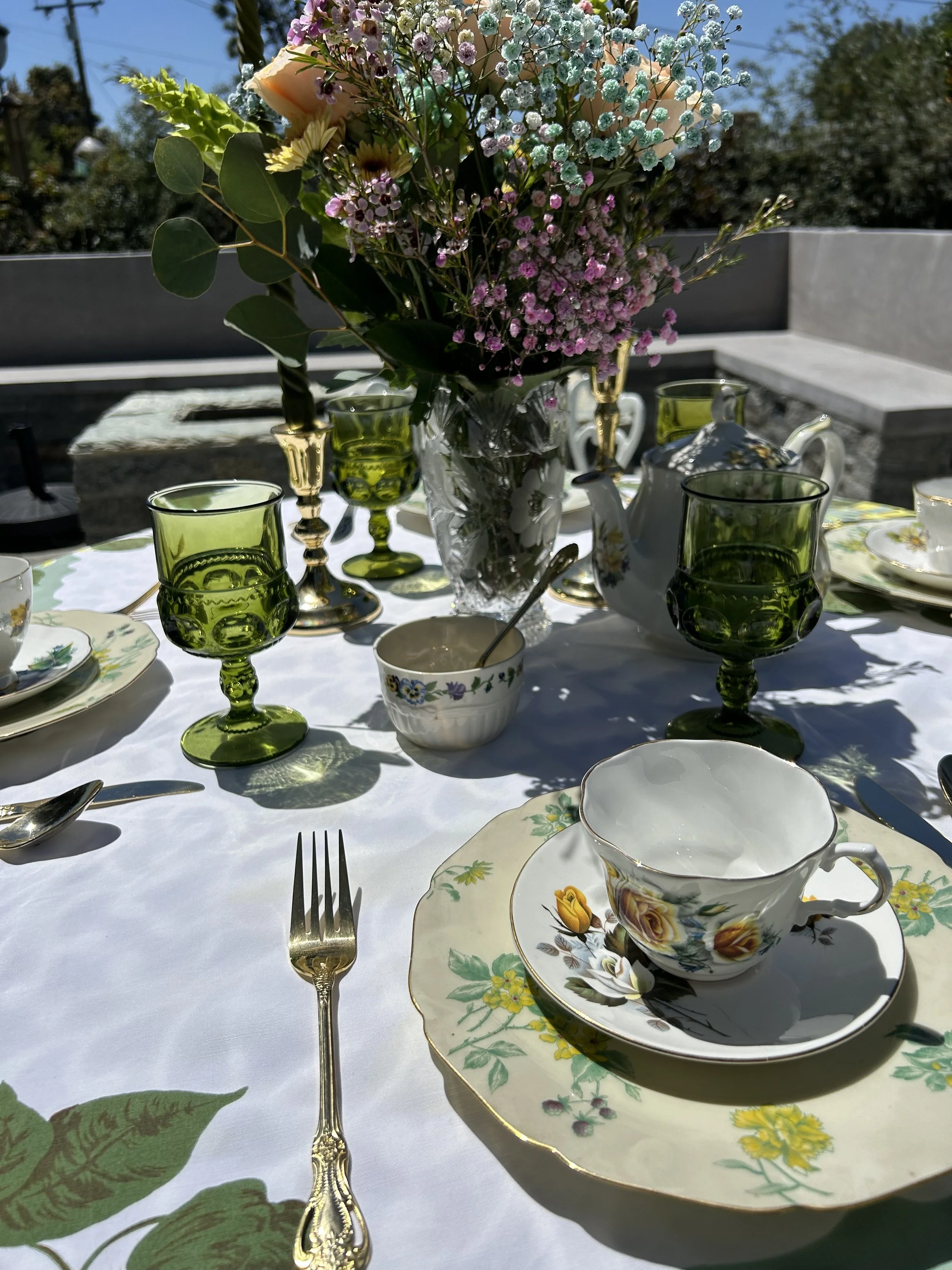 Outdoor table setting with floral china, green goblets, and a bouquet of flowers on a white tablecloth. Elegant cutlery and a teapot are also visible.