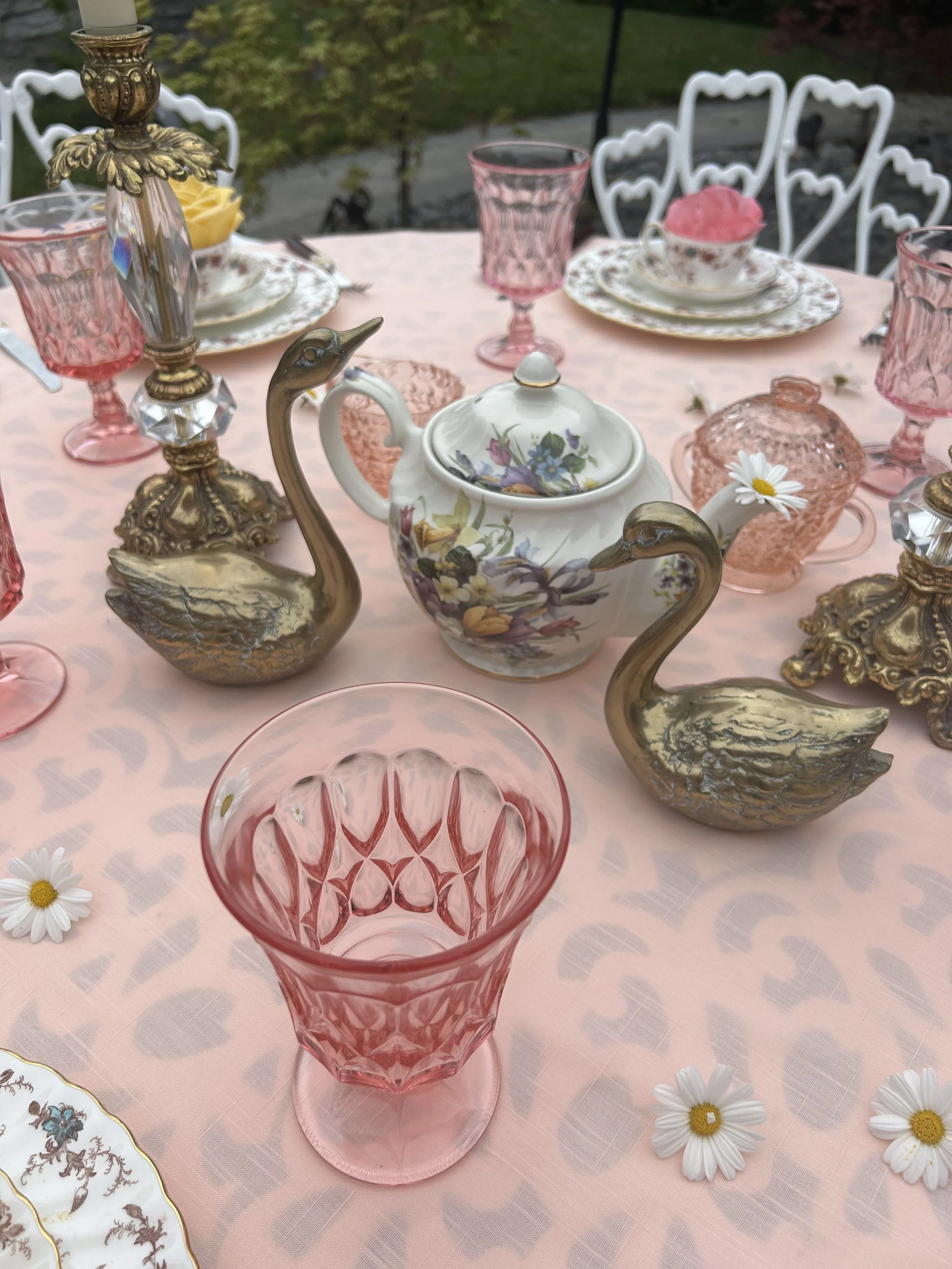 Elegant outdoor table setting with pink glassware, a floral teapot, ornamental brass swan figurines, and a floral-patterned teacup and saucer on a pink tablecloth. Daisy flowers are scattered on the table.