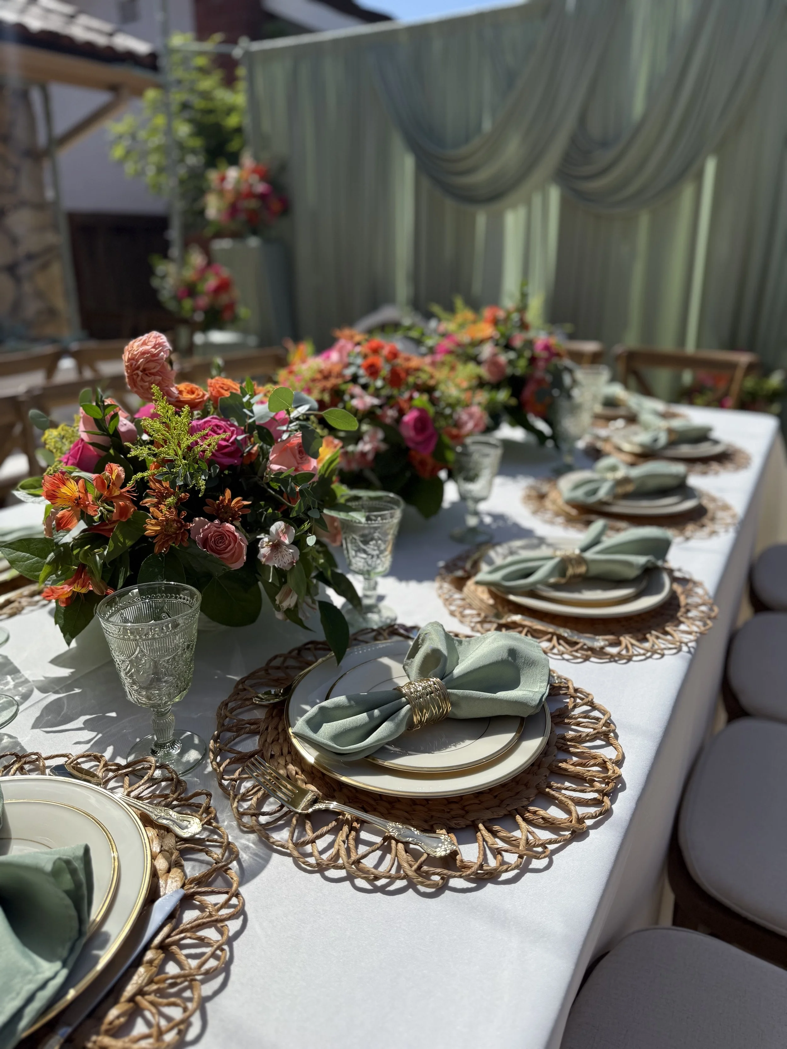 A beautifully decorated outdoor dining table with floral centerpieces, green napkins tied with gold rings on white plates, and crystal glasses, set for a special occasion with sunlight and a green curtain backdrop.