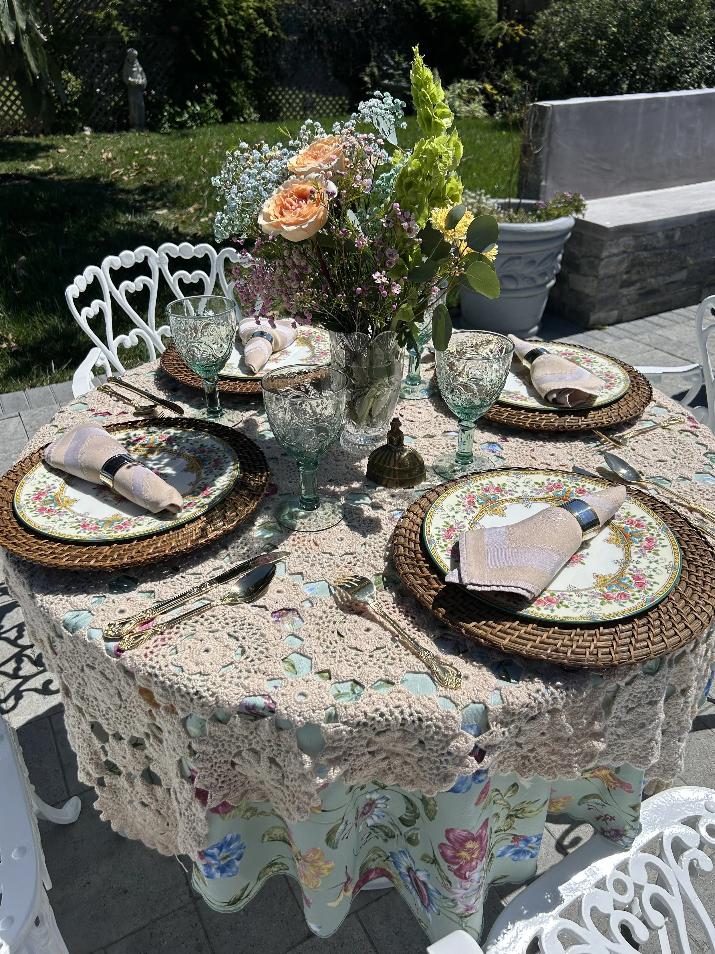 Outdoor table setting with floral decorations, lace tablecloth, vintage floral plates, woven chargers, and glassware.