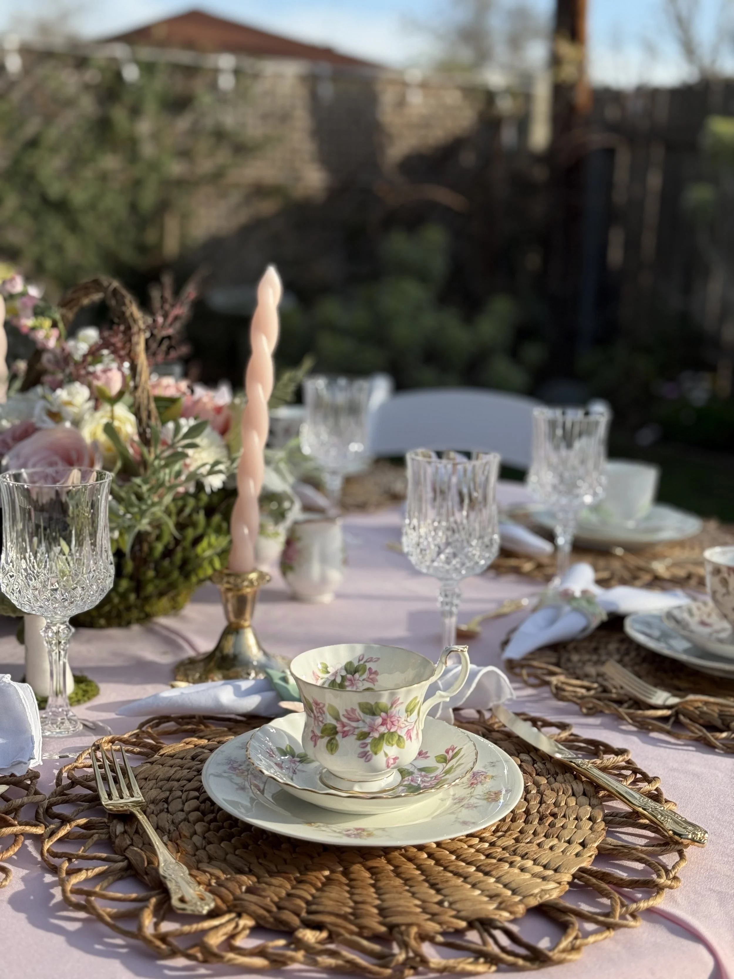 Table set for an outdoor meal with floral china tea cup and saucer on woven placemat, crystal glassware, silver fork and knife, pink candle, and floral centerpiece, in a garden with a stone wall and trees in the background.