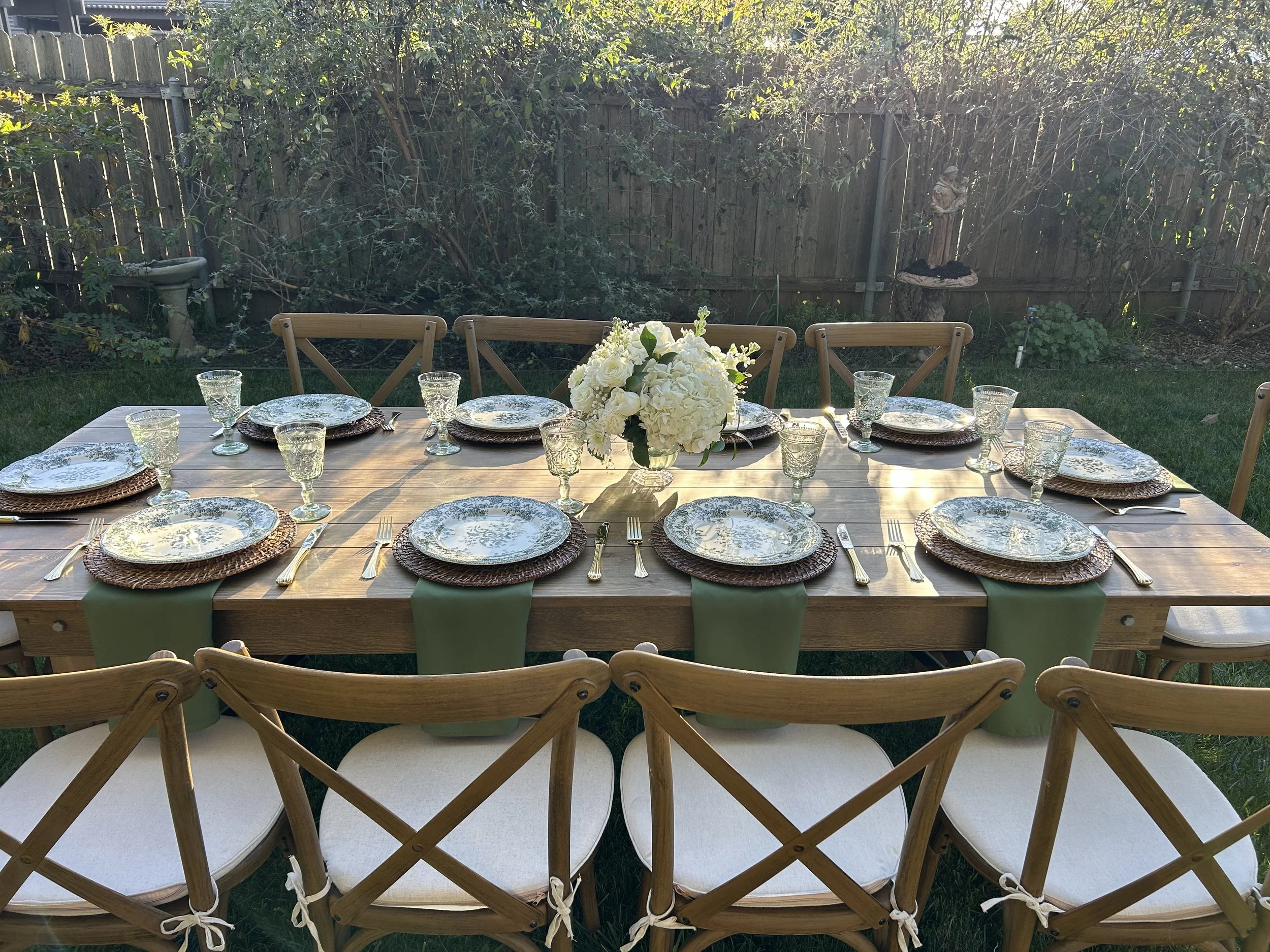 A wooden outdoor dining table set for a meal with eight place settings, each with a plate, gold-colored cutlery, and a green napkin, in a garden with a wooden fence and lush greenery in the background, and a floral centerpiece.