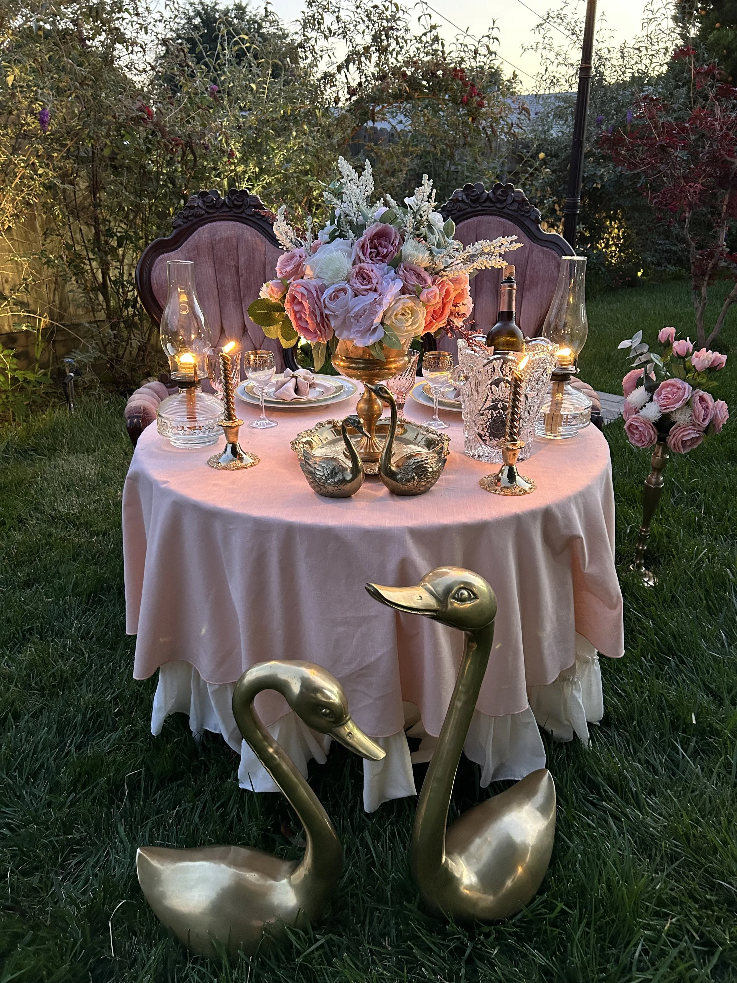 Outdoor romantic dinner setup with a round table draped in a soft pink tablecloth, adorned with flowers, vintage-style chairs, candles, a dinner setting, and decorative swan figurines on the grass.