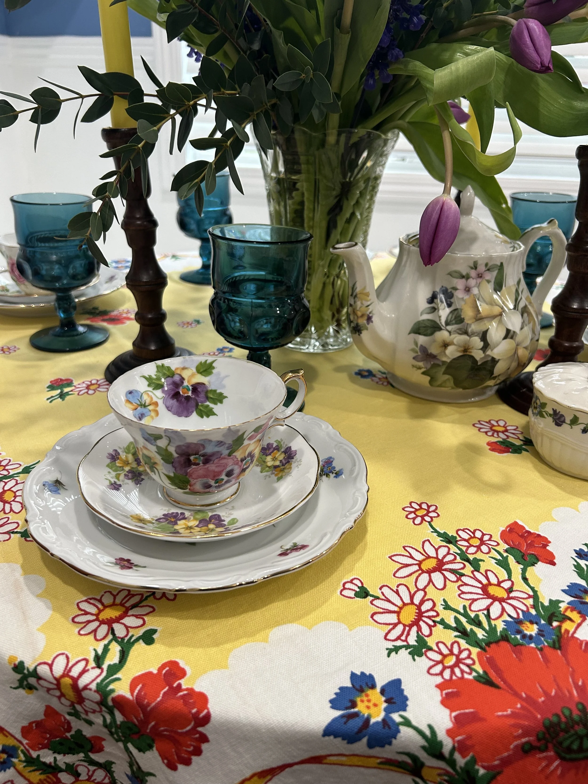 Table setting with floral tea set, blue glass goblets, bouquet of purple tulips, and floral tablecloth.