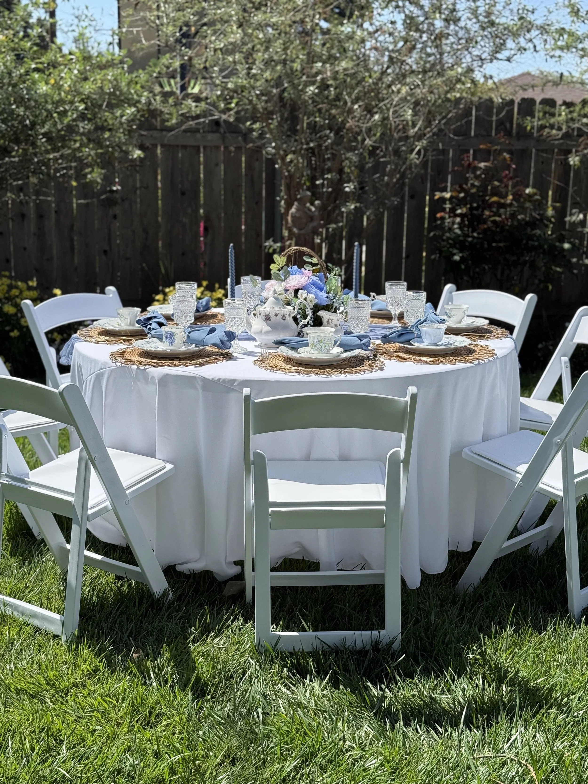Round outdoor dining table set with white tablecloth, blue napkins, and elegant tableware, surrounded by white folding chairs, in a garden with trees and a wooden fence.