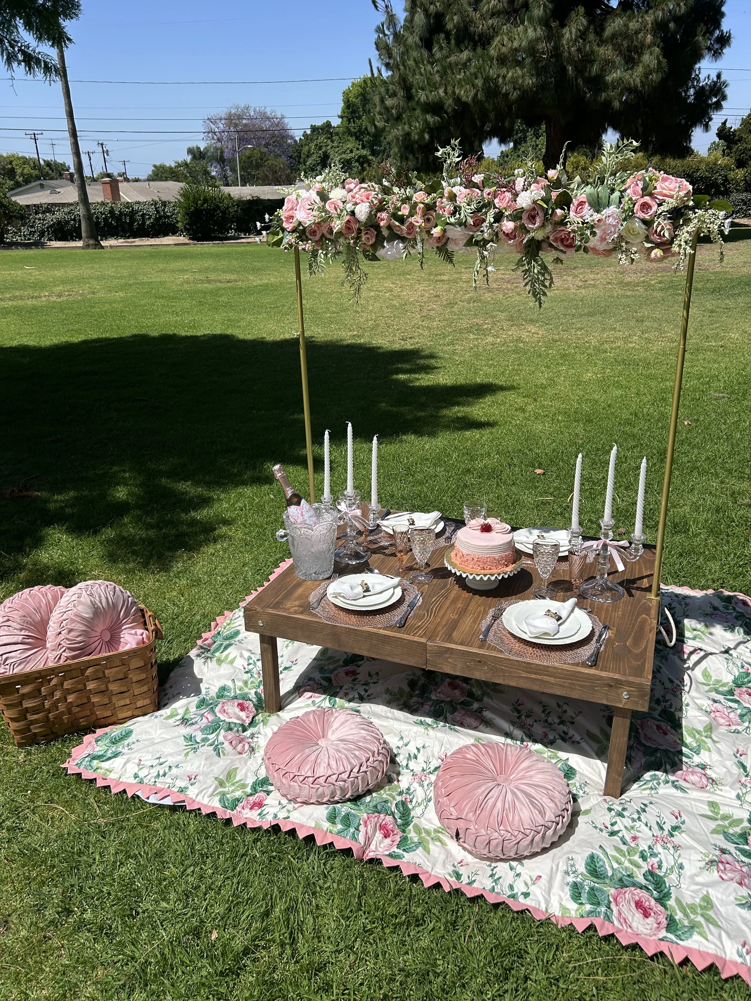 Outdoor picnic setup with a wooden table on a floral blanket, pink cushions, and a floral canopy. The table is set with plates, glasses, a pink cake, and candlesticks.