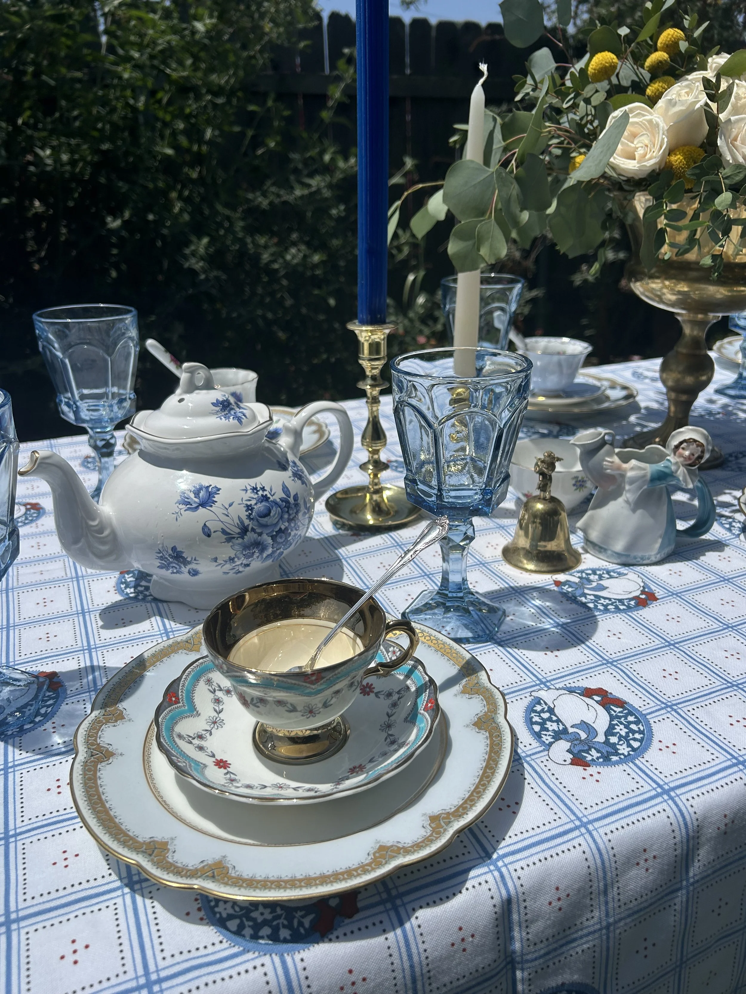 A vintage tea party setup featuring a floral teapot, gold-rimmed teacup, fine china plates, blue glassware, and brass candlesticks on a patterned tablecloth. The table is adorned with fresh flowers in a vase and decorative accessories.