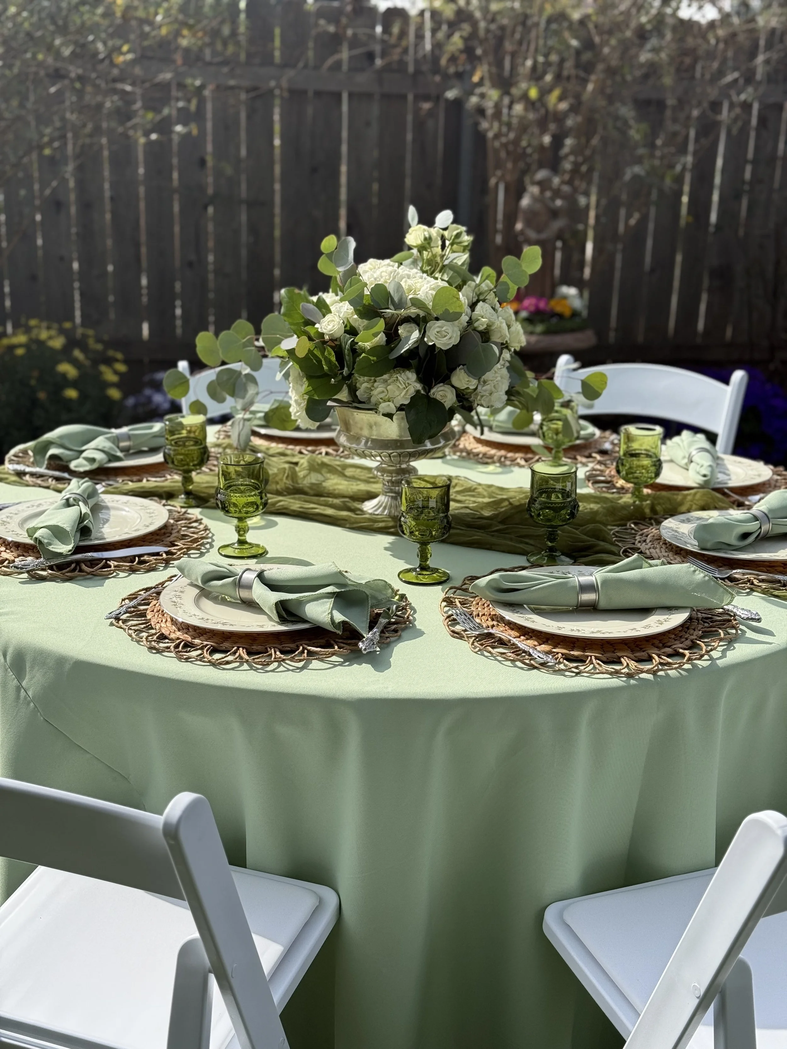 Table set for outdoor dining with green tablecloth, floral centerpiece, woven placemats, green glassware, and folded napkins.
