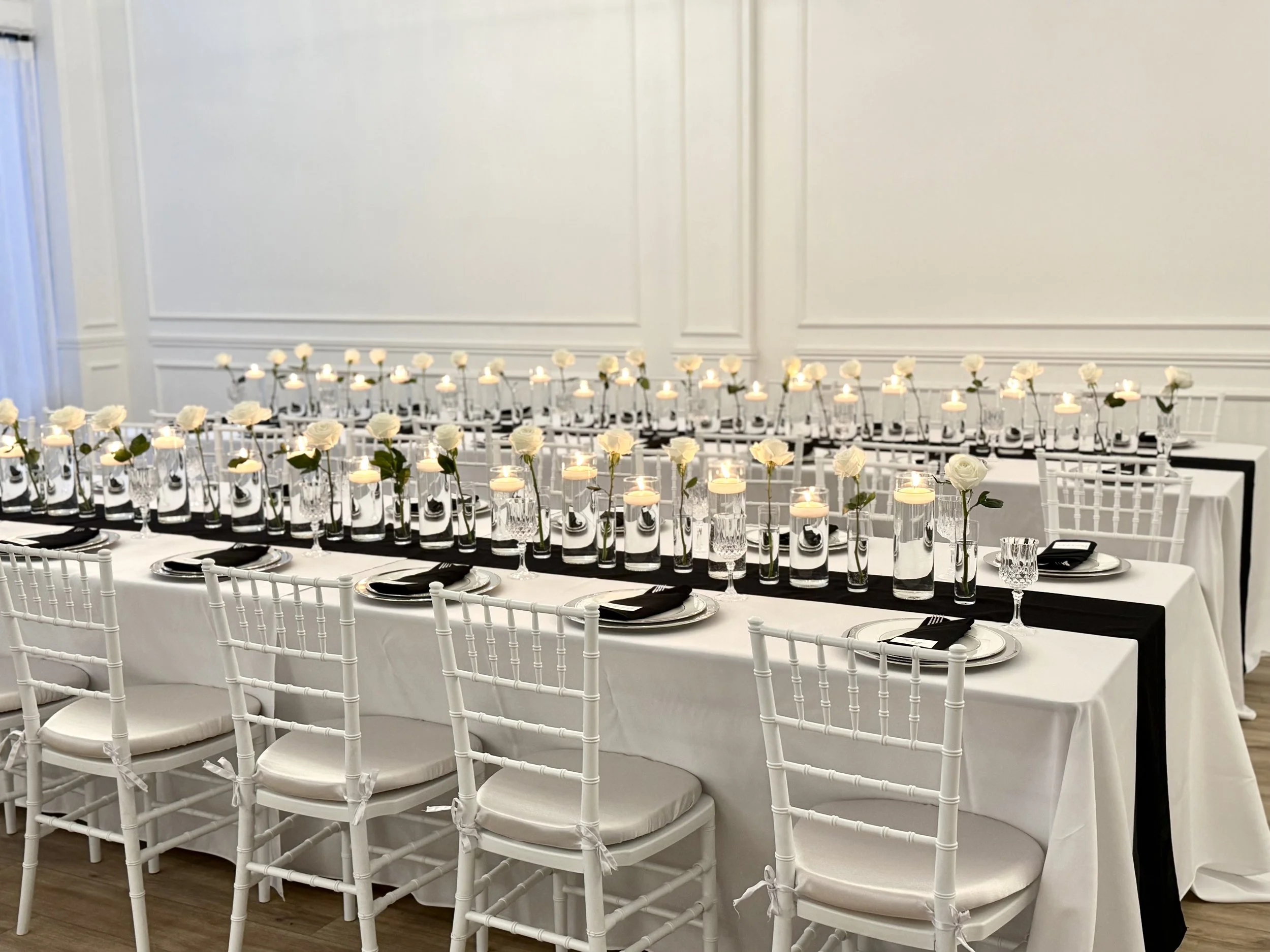Elegant dining table with white tablecloth, black napkins, white chairs, glassware, and centerpieces with white roses and candles in a banquet hall.