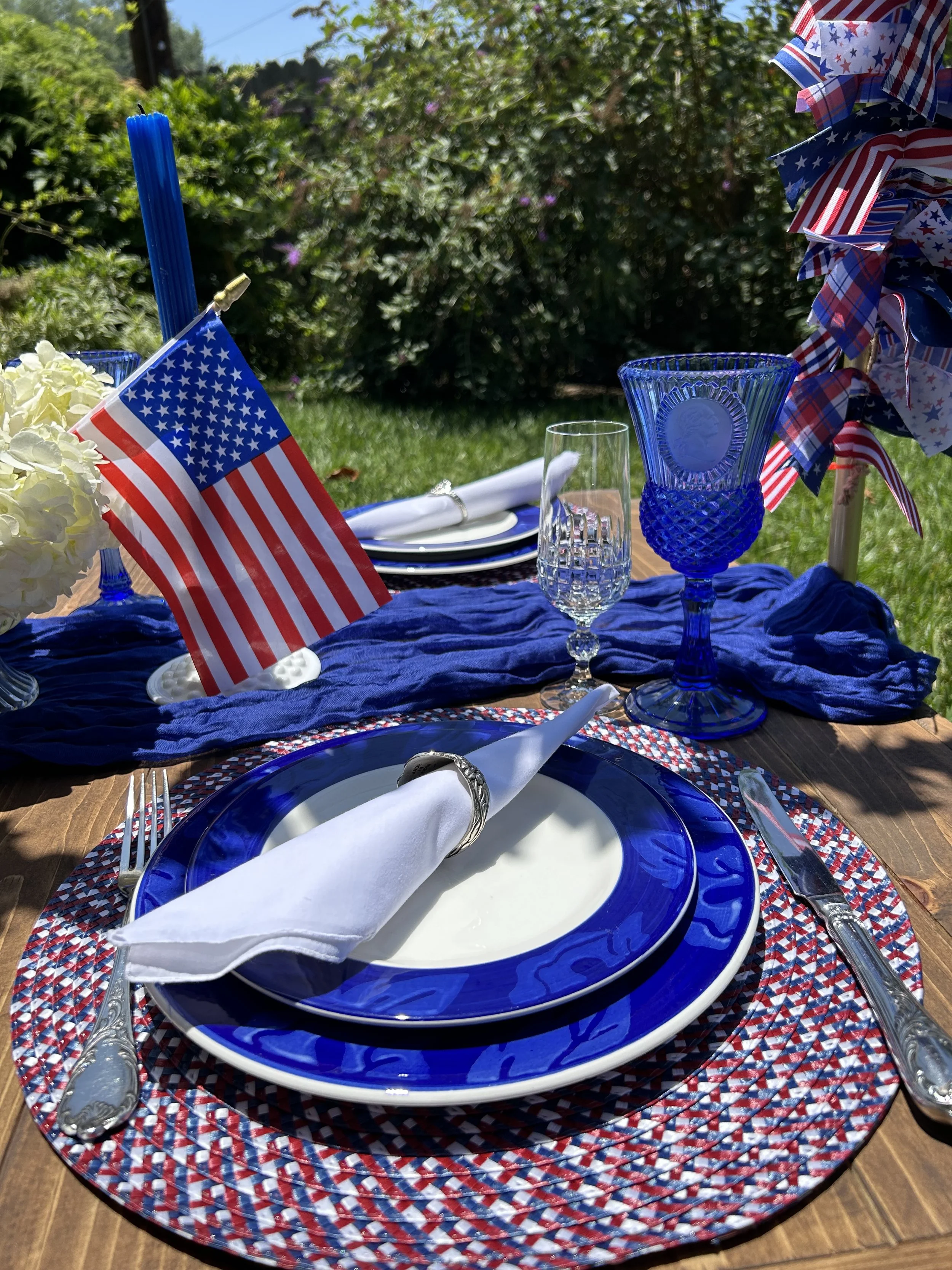 Patriotic outdoor table setting with American flag, blue and white plates, silverware, blue glassware, and red, white, and blue decorations on a wooden table.