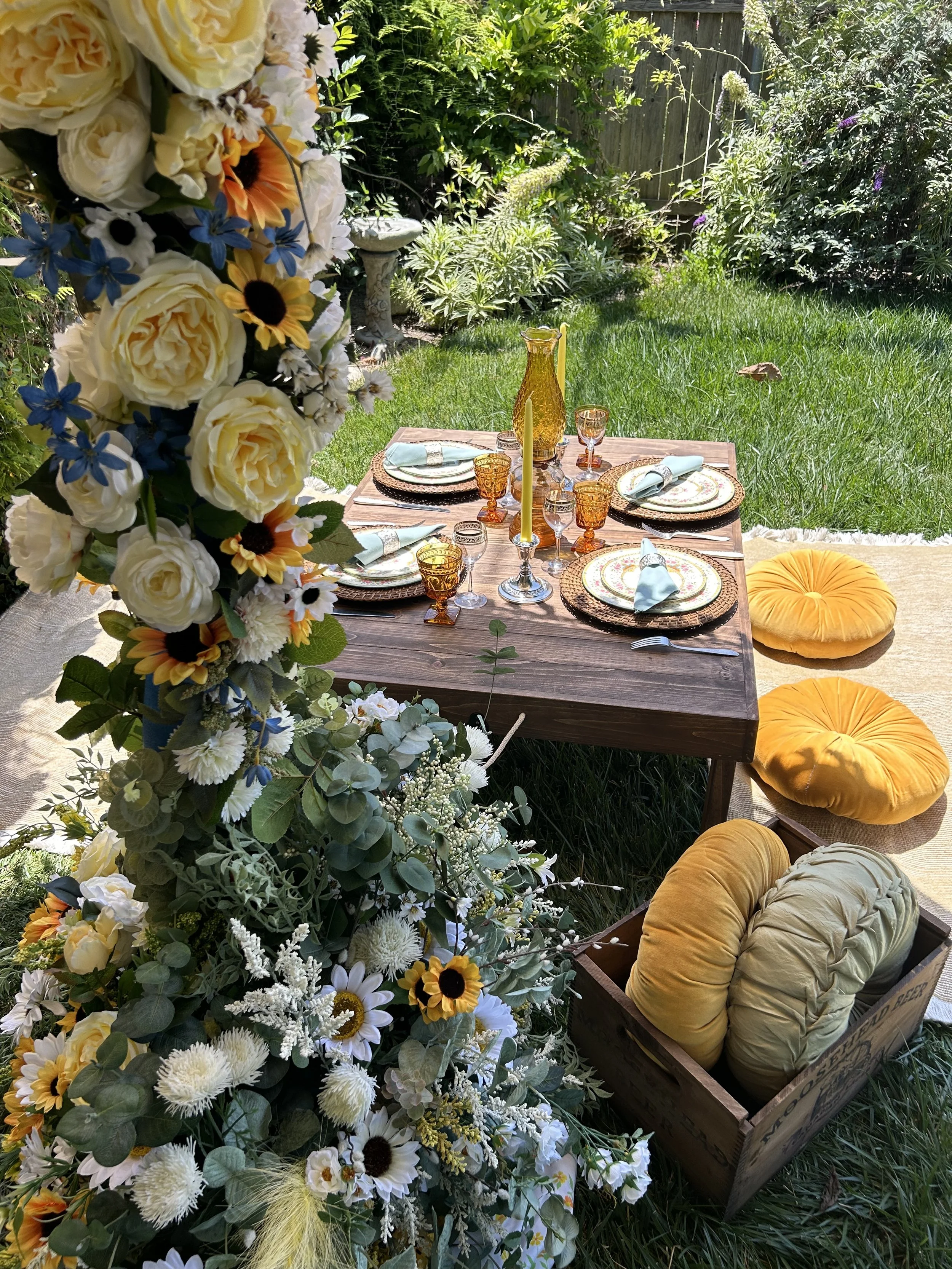 Outdoor table setting with floral arrangement, featuring yellow and white flowers and greenery. The wooden table has vintage-style glassware, blue napkins, and decorative plates. Yellow cushions and a box of round pillows are nearby, situated on a grassy lawn surrounded by plants.