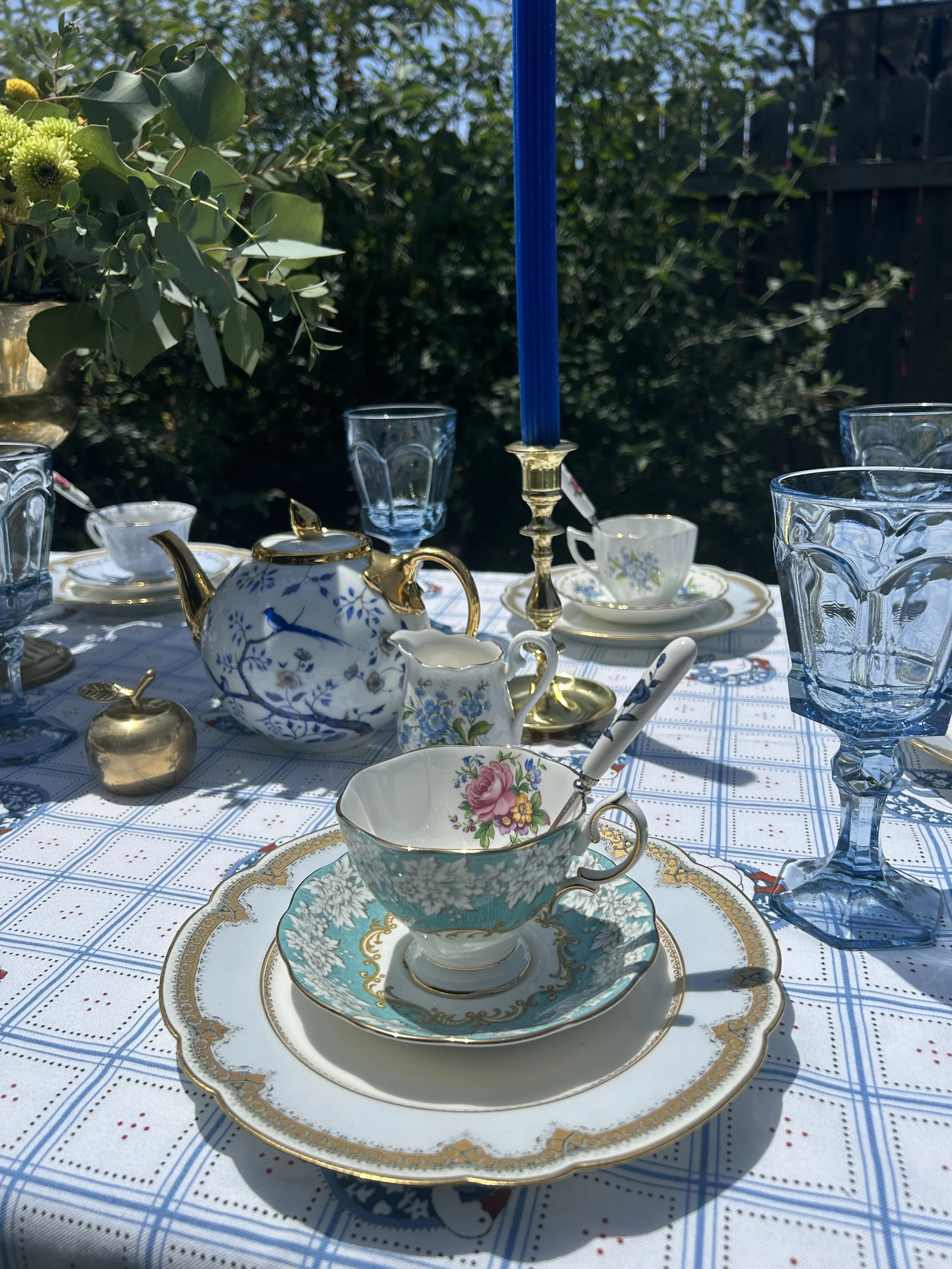 Elegant outdoor table setting with floral teacups, blue glassware, gold candle holders, a teapot, and a floral tablecloth.