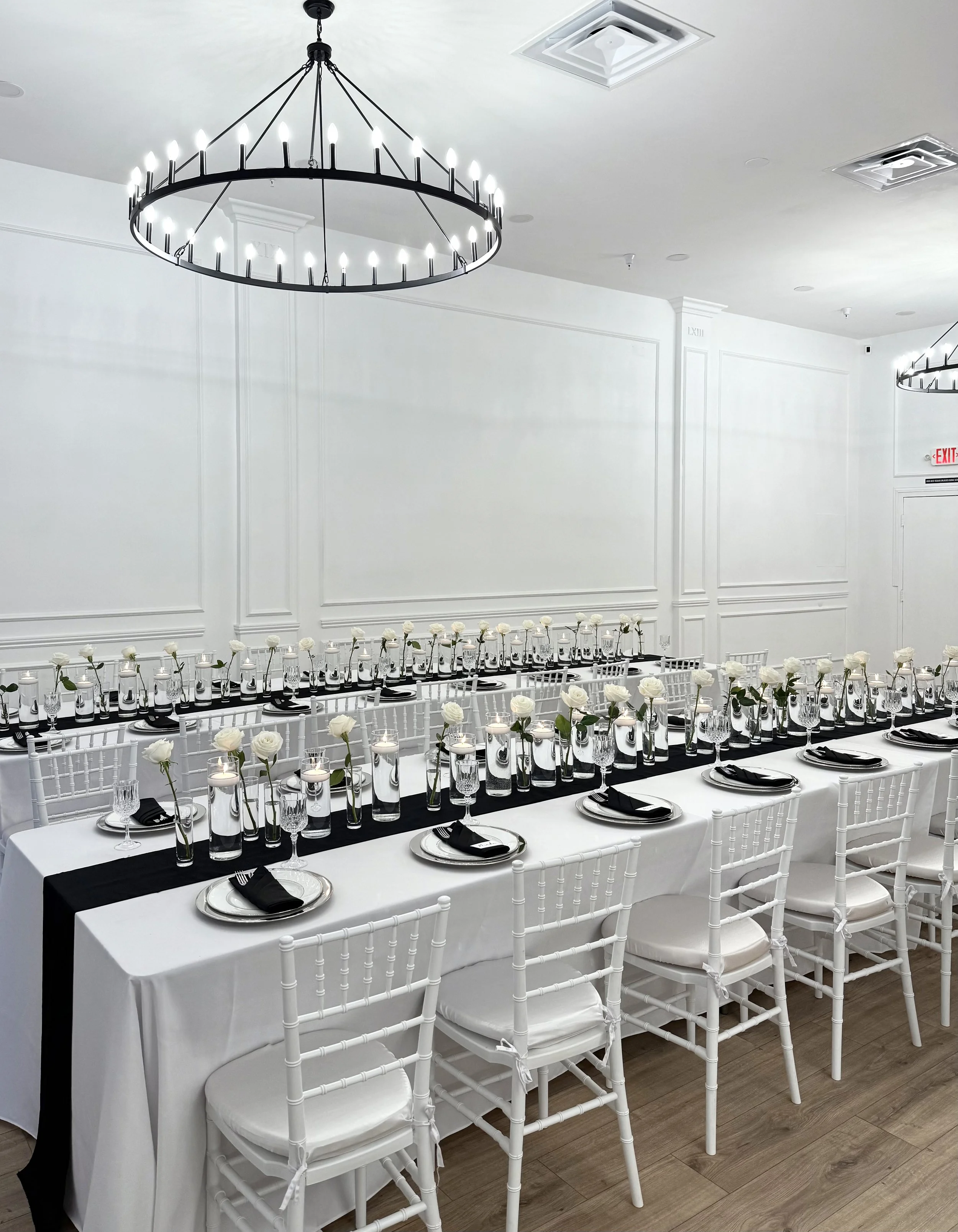 Elegant banquet table set with black and white decor, featuring white chairs, white tablecloths, black napkins, and white roses in tall vases, with a modern chandelier overhead in a white room.