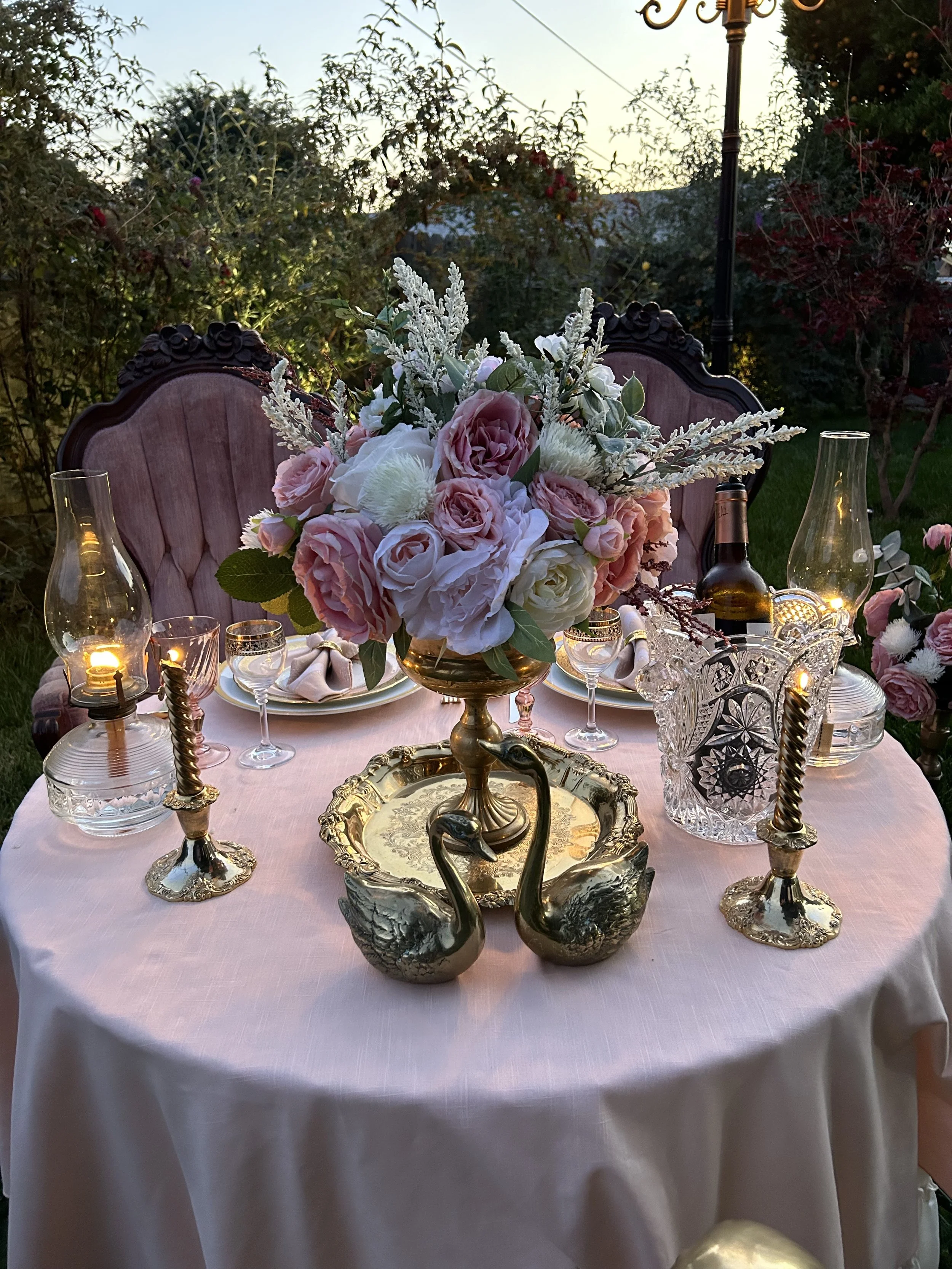 Elegant outdoor dining setup with pink tablecloth, floral centerpiece, vintage swan decor, candlesticks, glassware, and oil lamps.