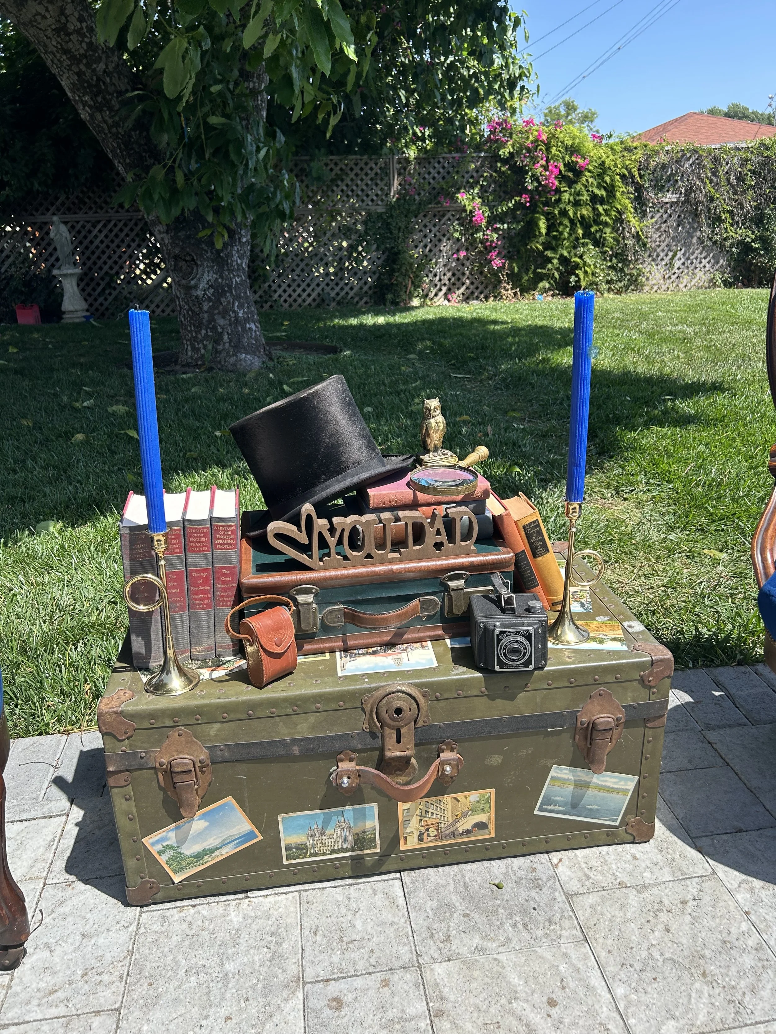 Vintage setup with an old trunk decorated with postcards, topped with books, a top hat, camera, "You're Dad" sign, owl figurine, blue candles, and brass horns, set against a garden backdrop.