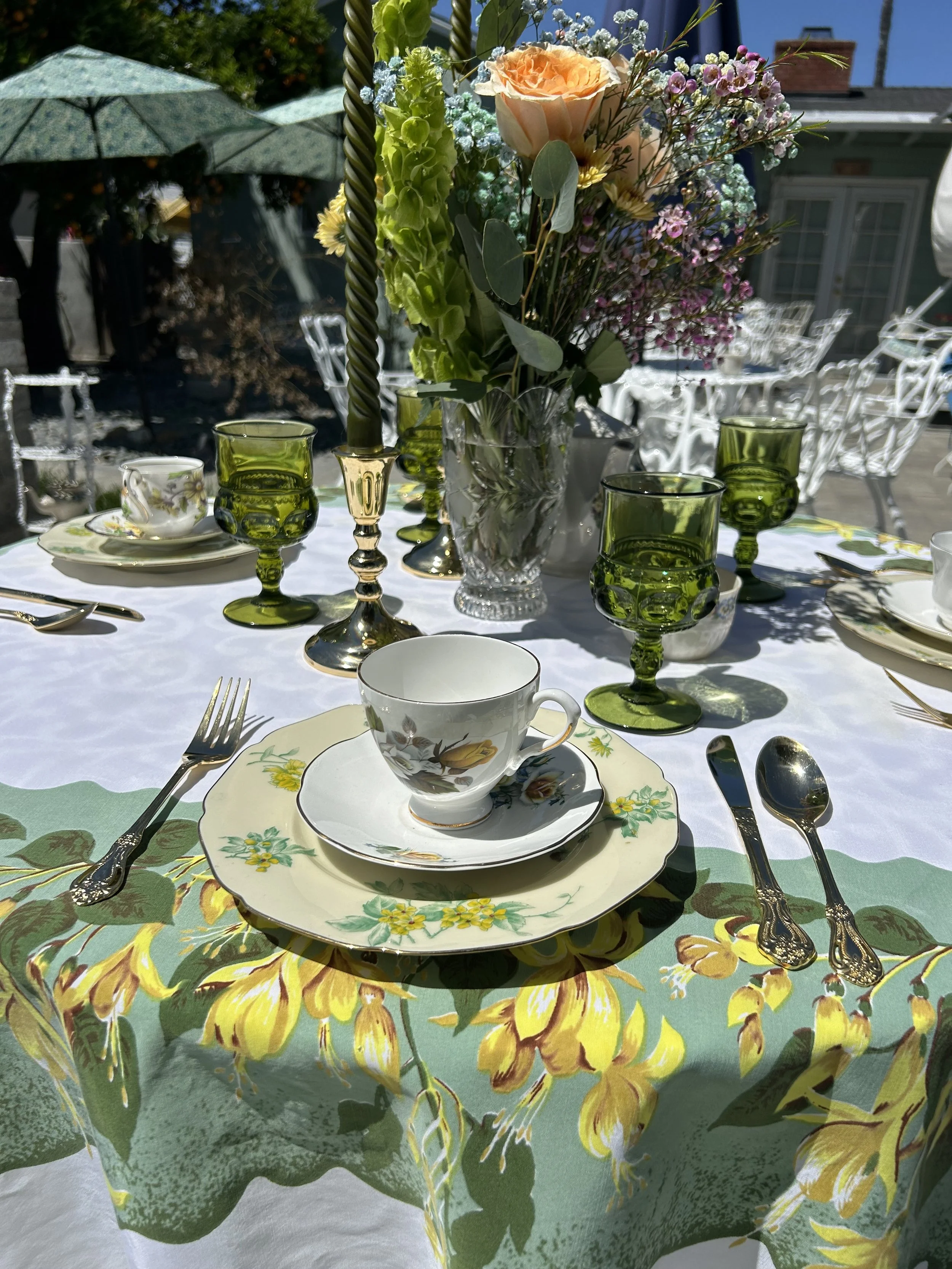 Outdoor dining table setting with floral tablecloth, green glassware, floral china, silverware, a vase of mixed flowers, and green candles.