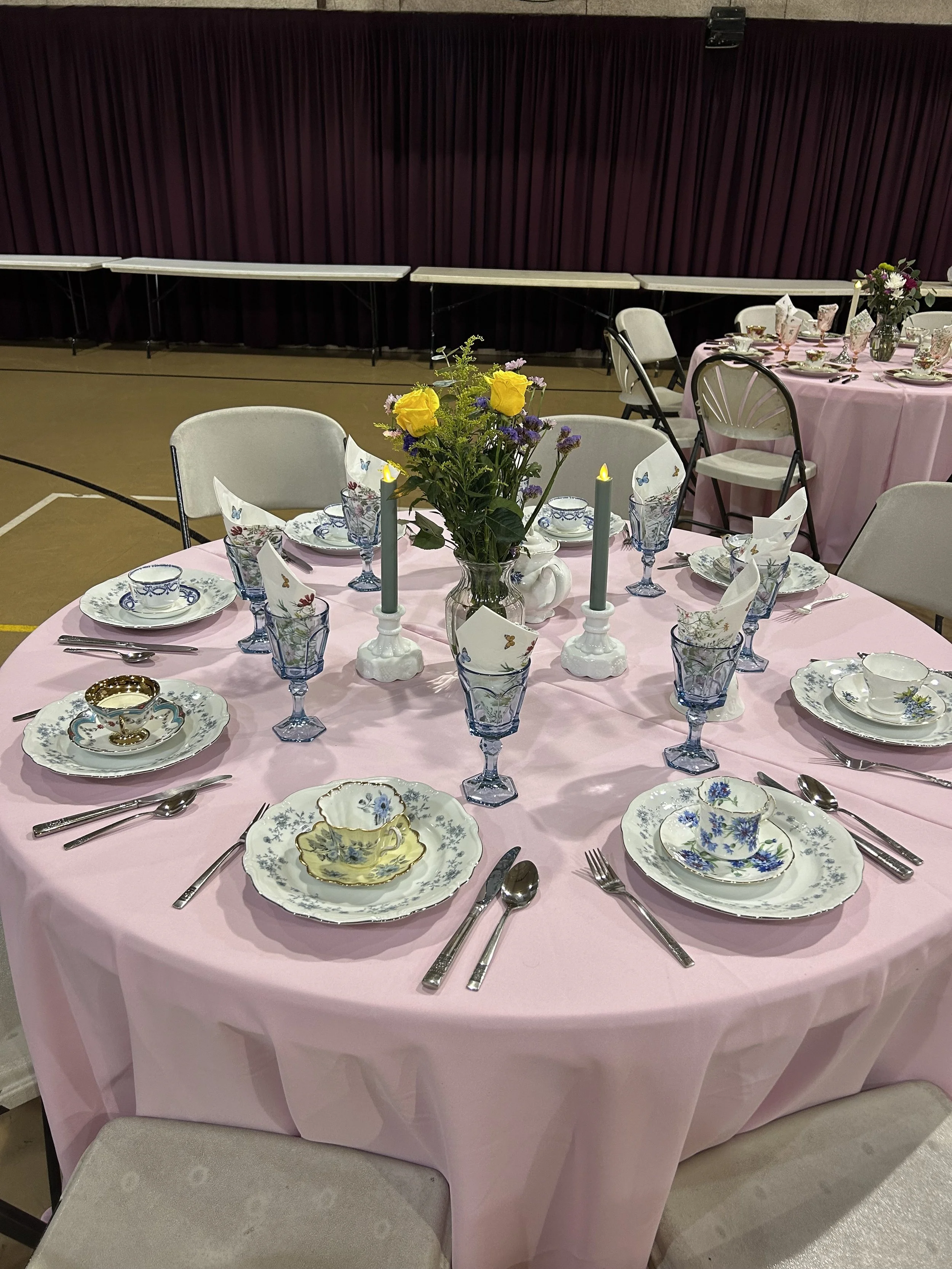 Table set with pink tablecloth, floral teacups, blue glasses, and silver utensils; centerpiece with yellow flowers and candles.