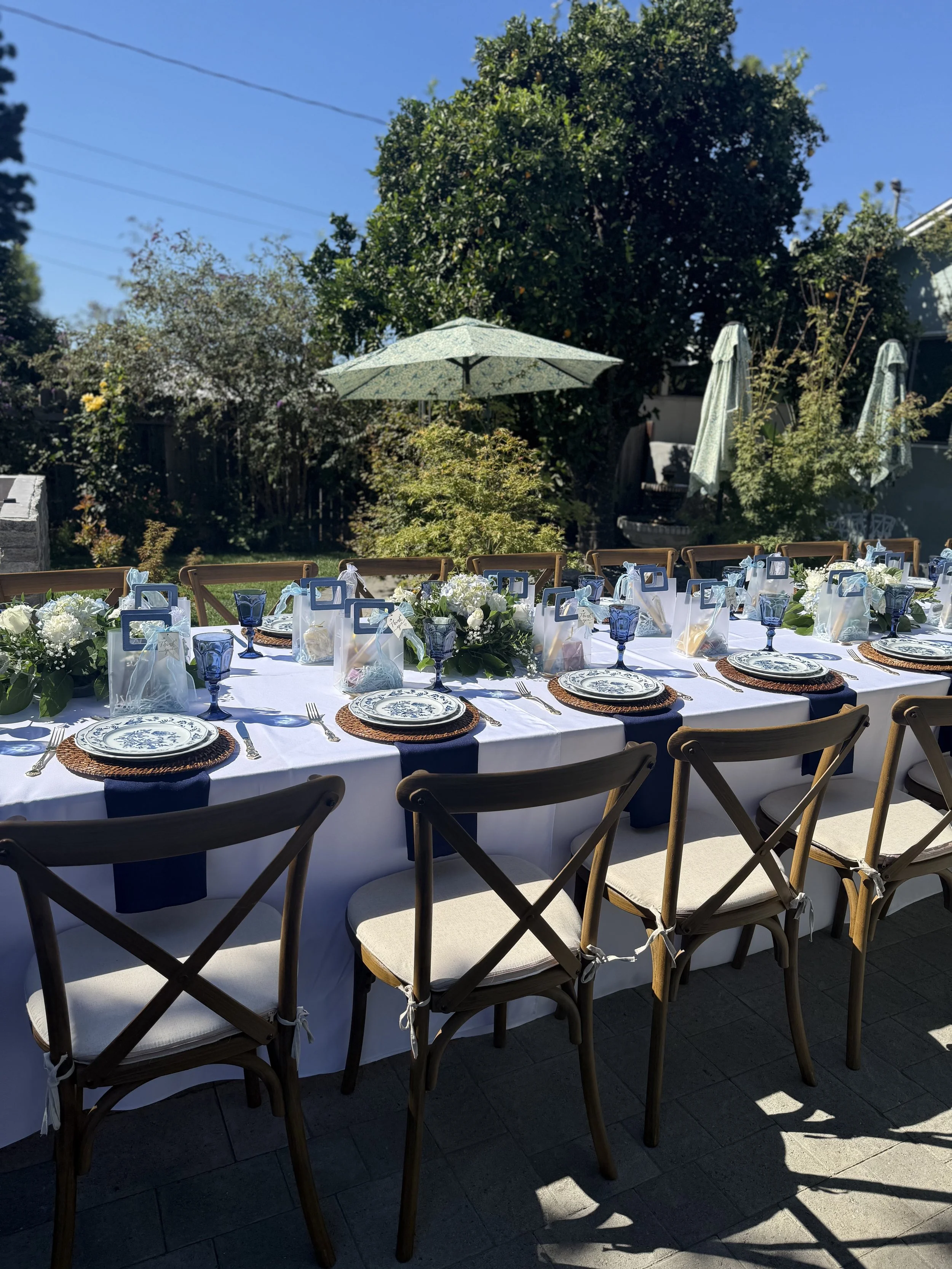 Outdoor dining table set for a celebration with white tablecloth, navy napkins, floral centerpieces, blue glasses, and decorative gift bags, in a backyard with green trees, umbrellas, and a clear blue sky.