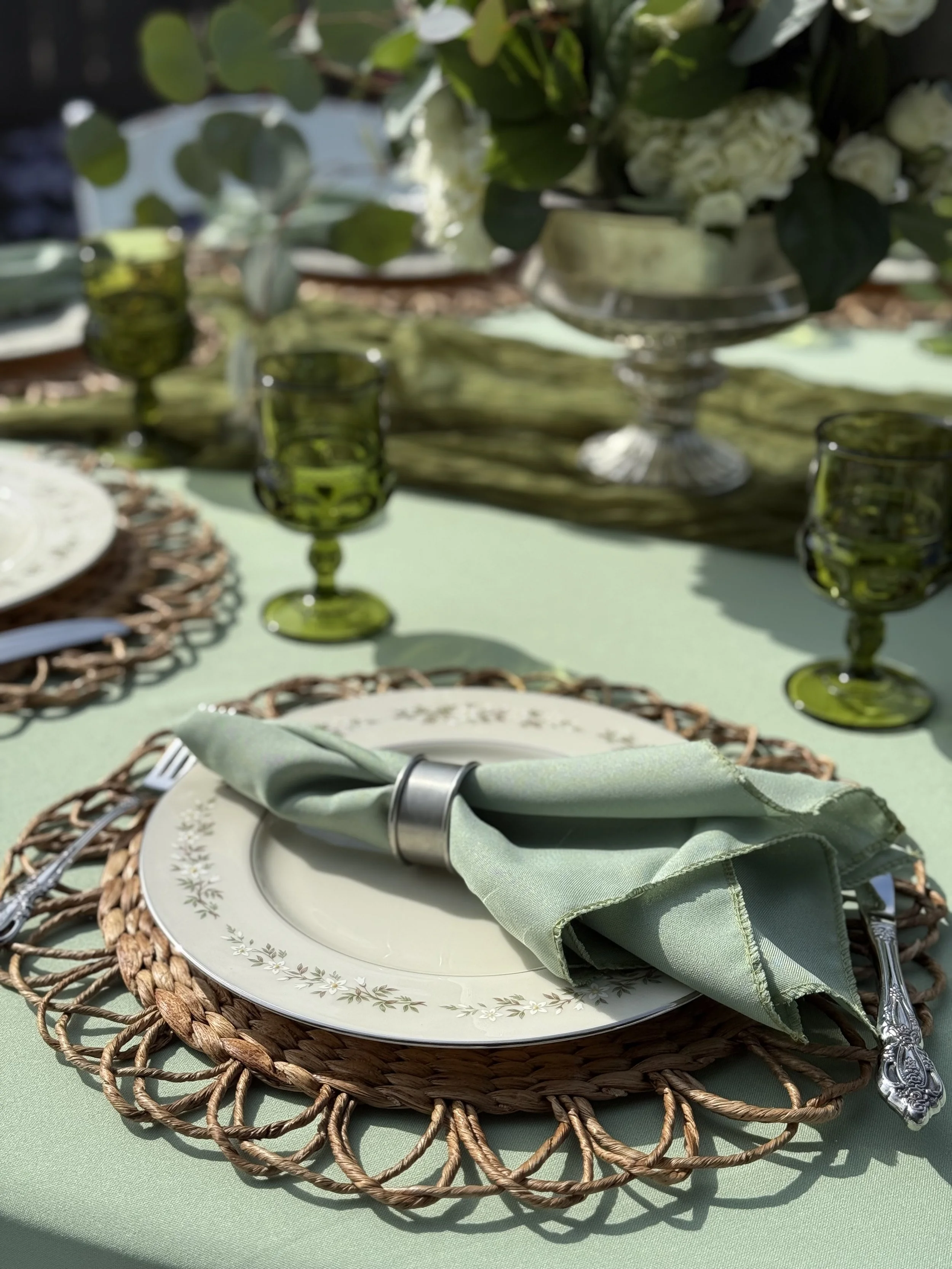 Elegant outdoor table setting with green tablecloth, woven placemat, decorative plate, folded green napkin with silver napkin ring, vintage-style cutlery, green glassware, and floral centerpiece.
