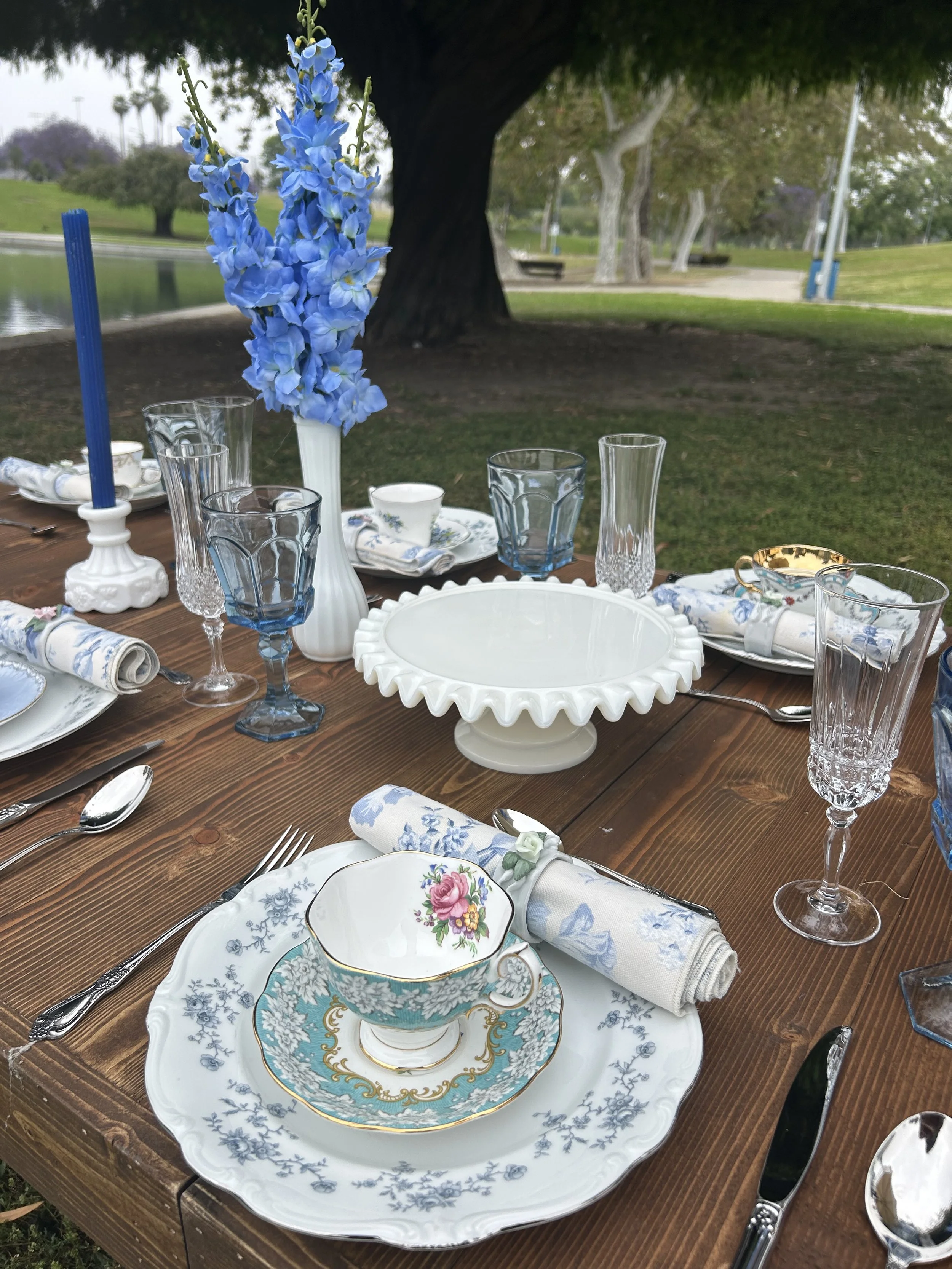 Outdoor table setting with vintage-style china, including a floral-patterned cup and saucer, silverware, and crystal glasses. A blue floral arrangement and blue candle in white holders are in the center. A wooden picnic table in a park setting with grass and trees in the background.