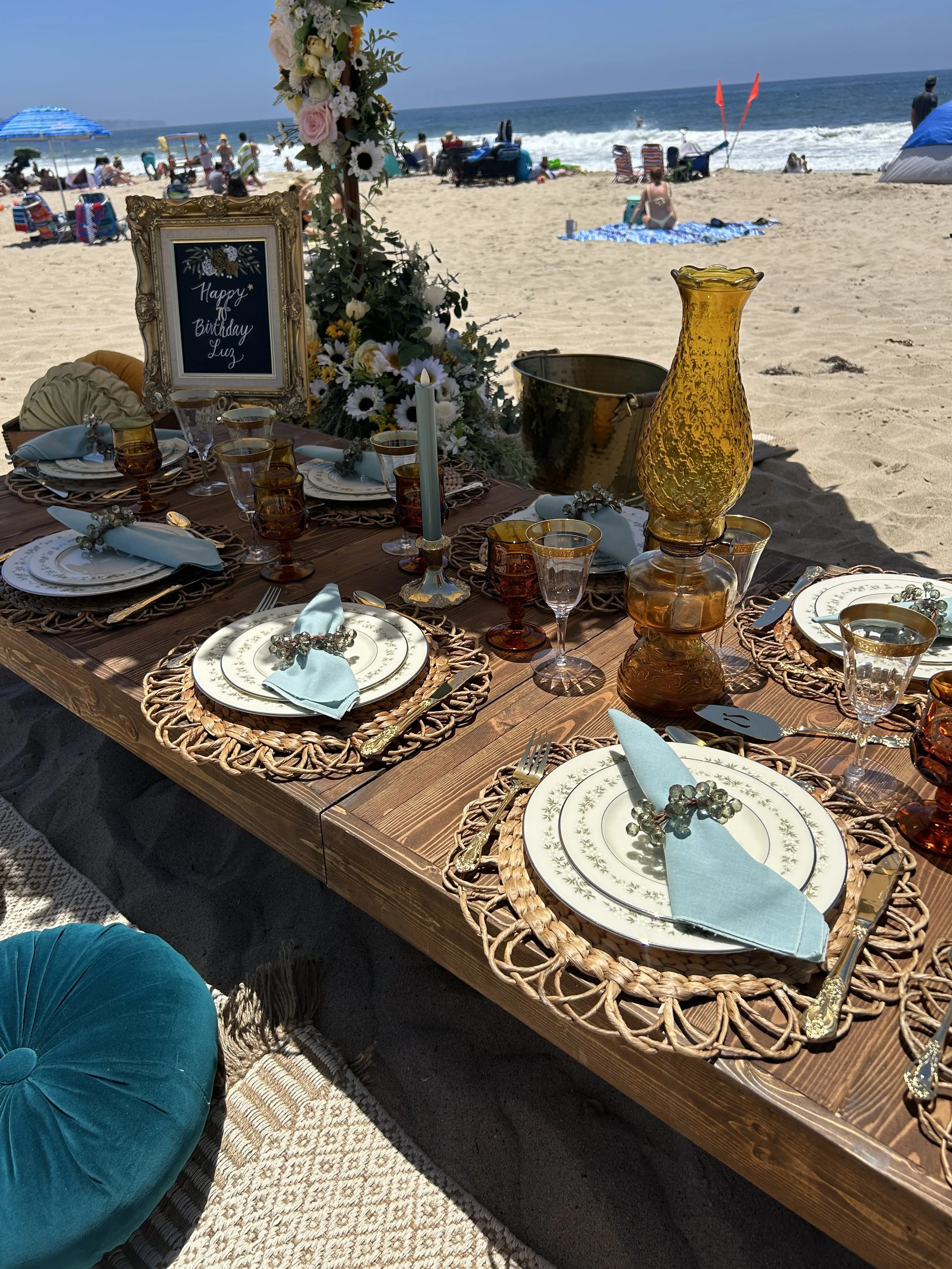 Beach picnic setup with a wooden table, decorative plates, blue napkins, amber glassware, and floral arrangements, set against a seaside backdrop with waves and people in the distance.