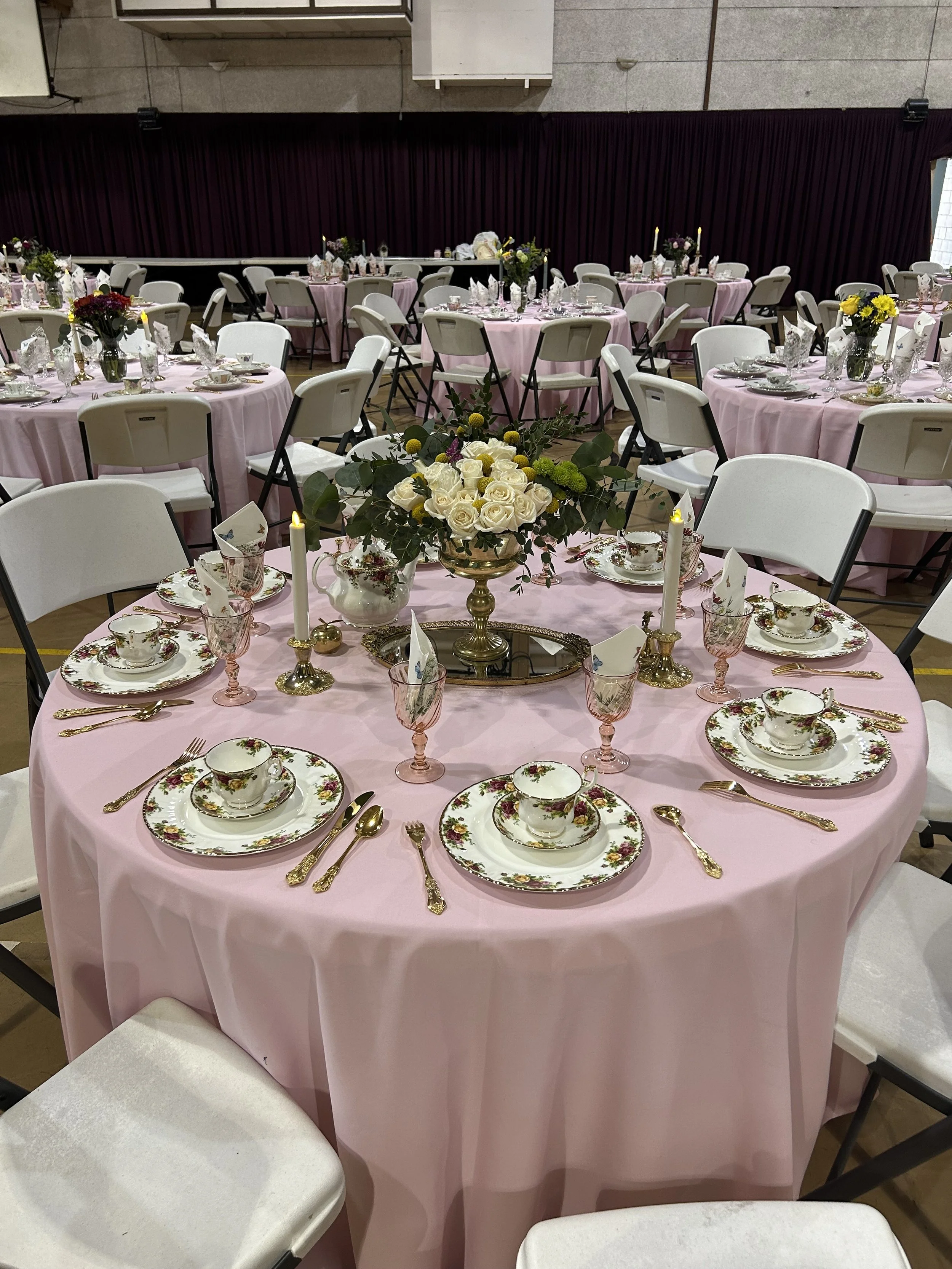 Ornately set round tables with pink tablecloths, white folding chairs, floral centerpieces, and floral-patterned china in a banquet hall setting.
