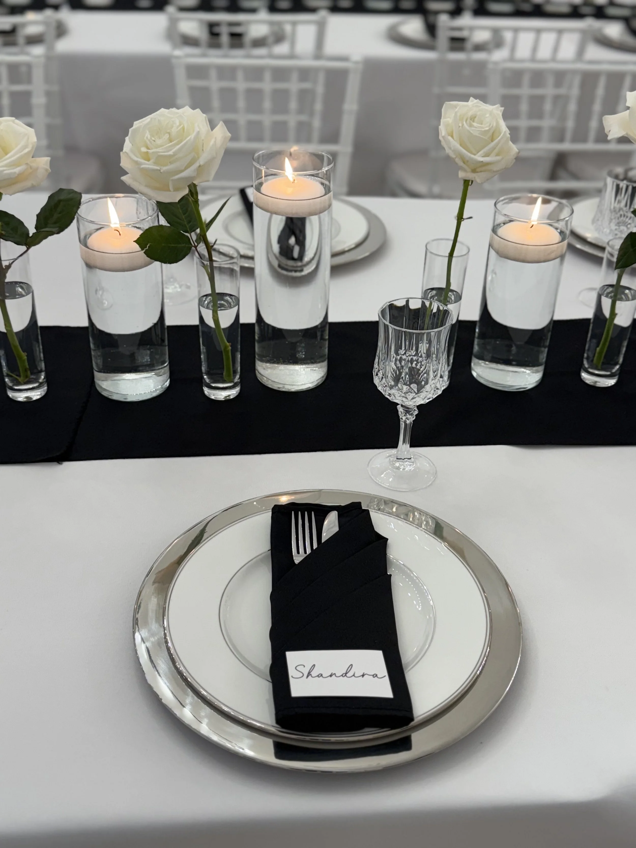 Elegant table setting with white roses in tall glass vases, lit candles, a crystal glass, a silver-rimmed plate, and a black napkin with a place card that reads 'Shandira'.