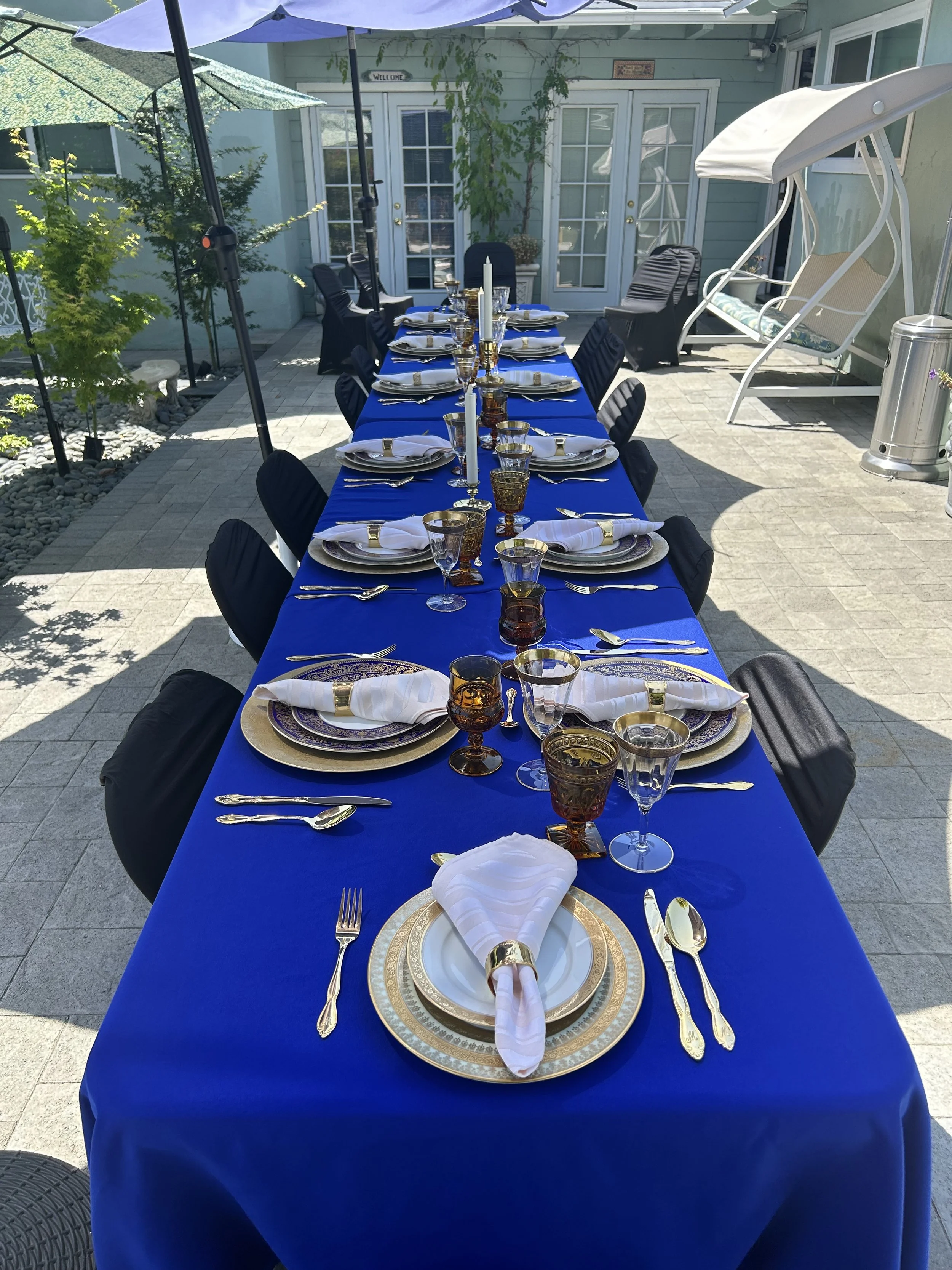 Outdoor dining table with blue tablecloth, set with elegant gold-trimmed plates, napkins, cutlery, and decorative glasses in a patio setting.