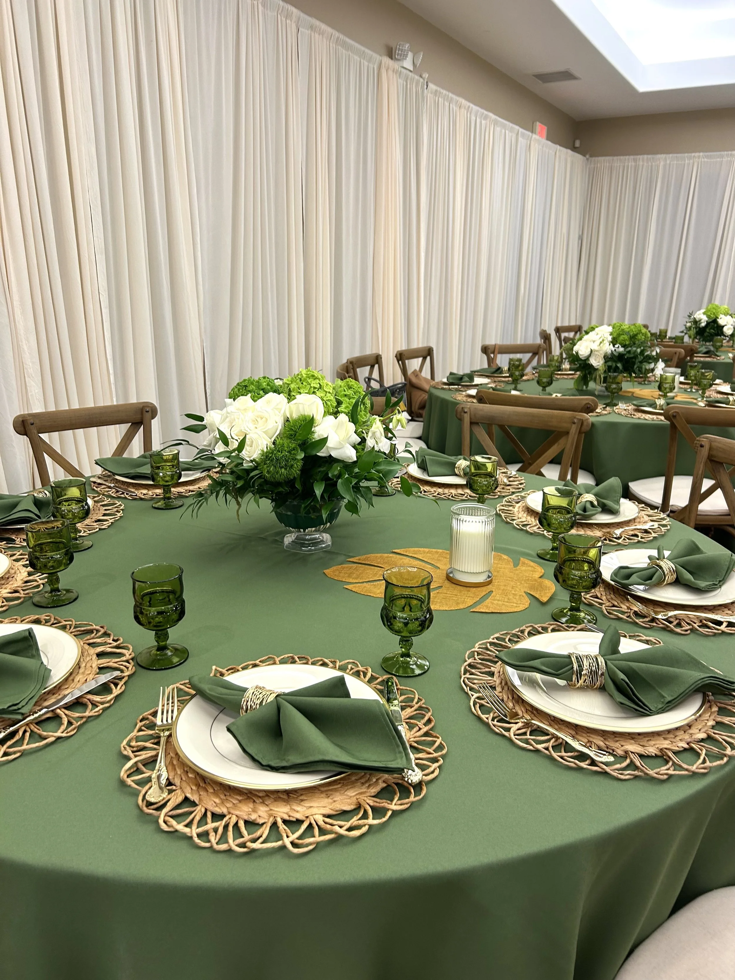 Elegant banquet table decorated with green tablecloth, green glassware, white plates with green napkins, floral centerpieces with white flowers and greenery, and candle decor in a room with white drapes.
