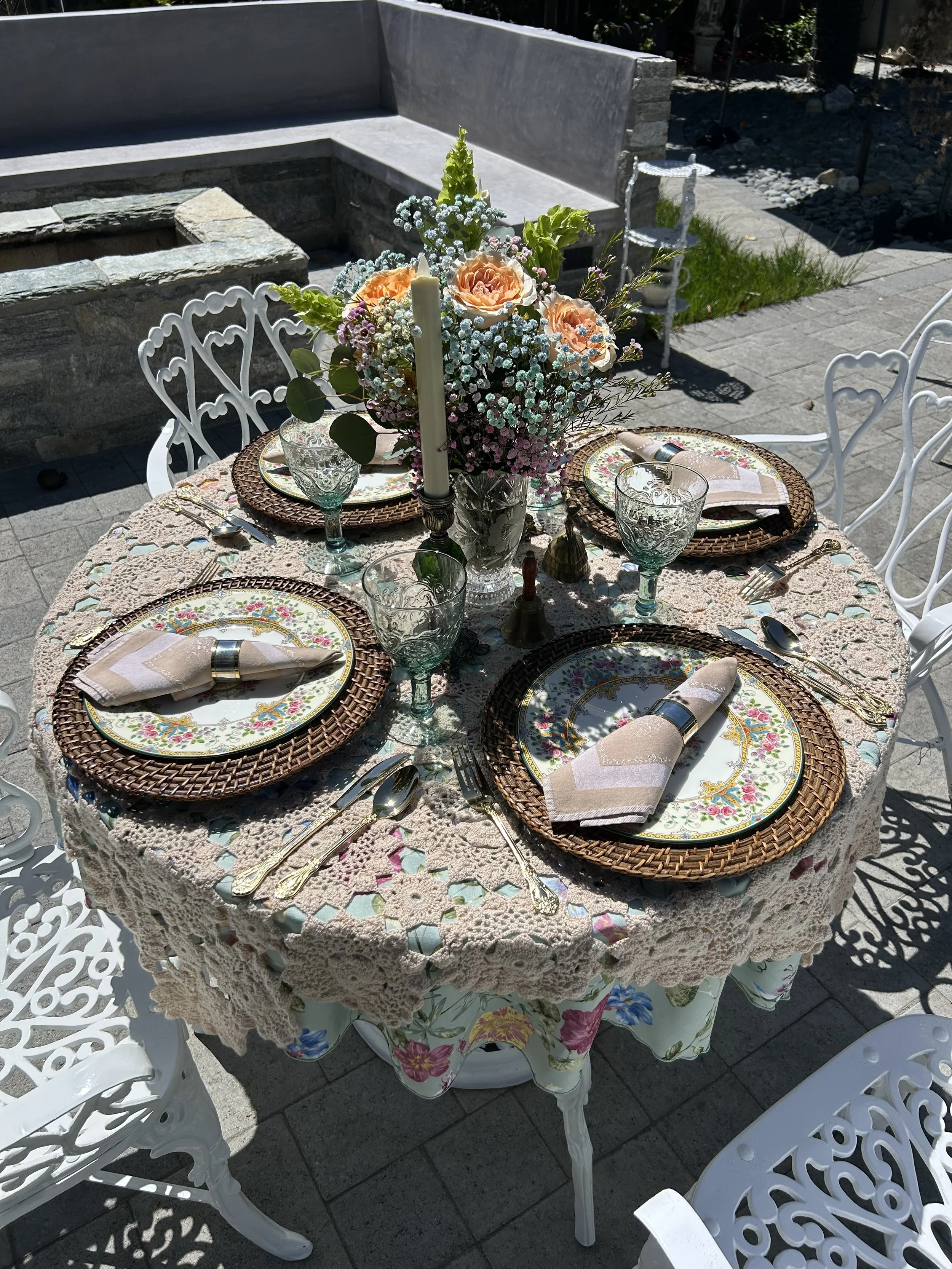 A beautifully set outdoor table with a floral tablecloth, lace overlay, and place settings for four. Each setting includes floral patterned plates, woven chargers, glass goblets, rolled napkins with napkin rings, and silverware. A large floral centerpiece with roses and baby’s breath is in the middle, accompanied by a candle. White ornate chairs surround the table on a stone patio.