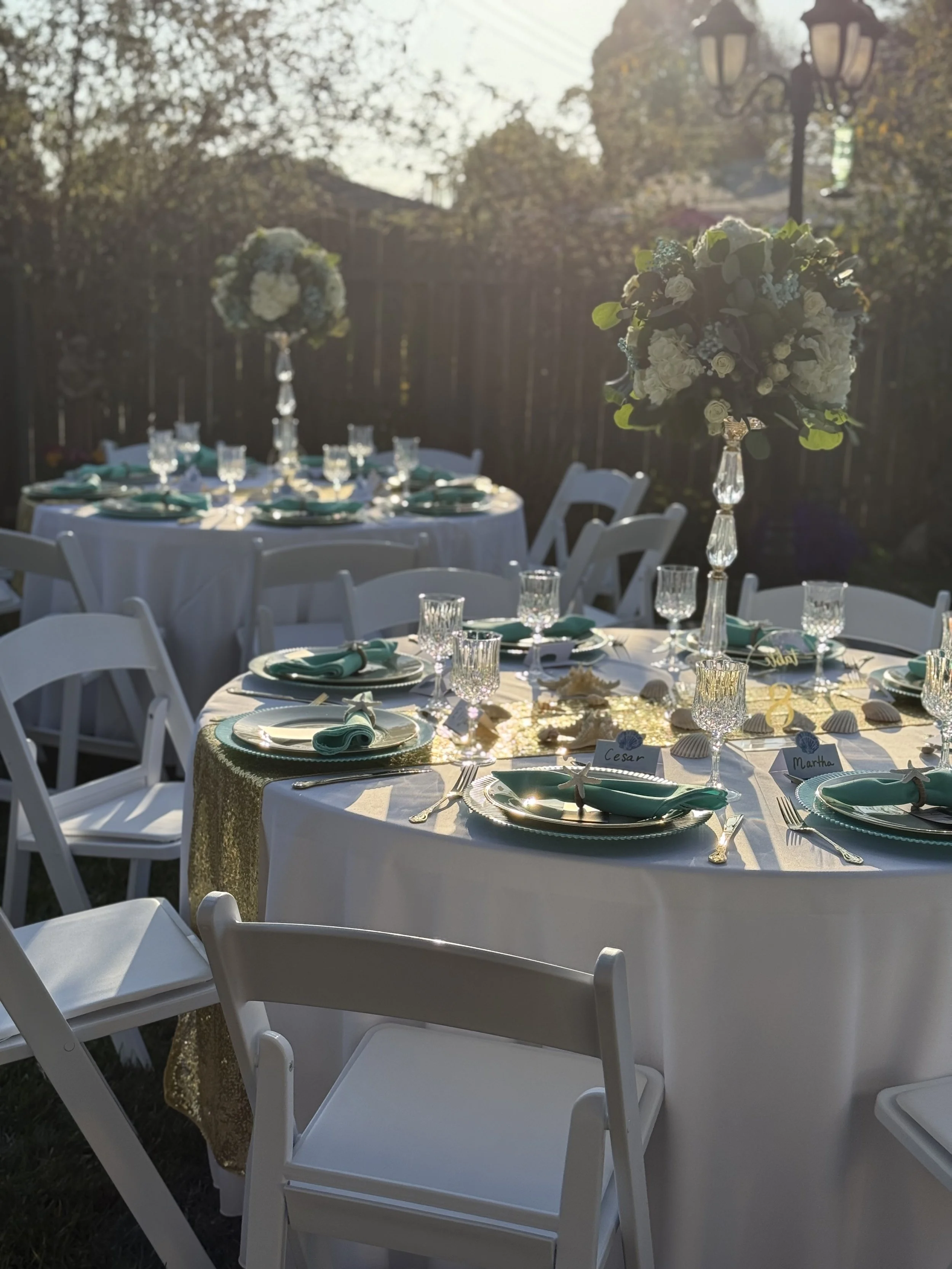 Outdoor table setup for event with white chairs, white tablecloth, teal plates, and glassware. Floral centerpieces on each table. Event under natural sunlight.