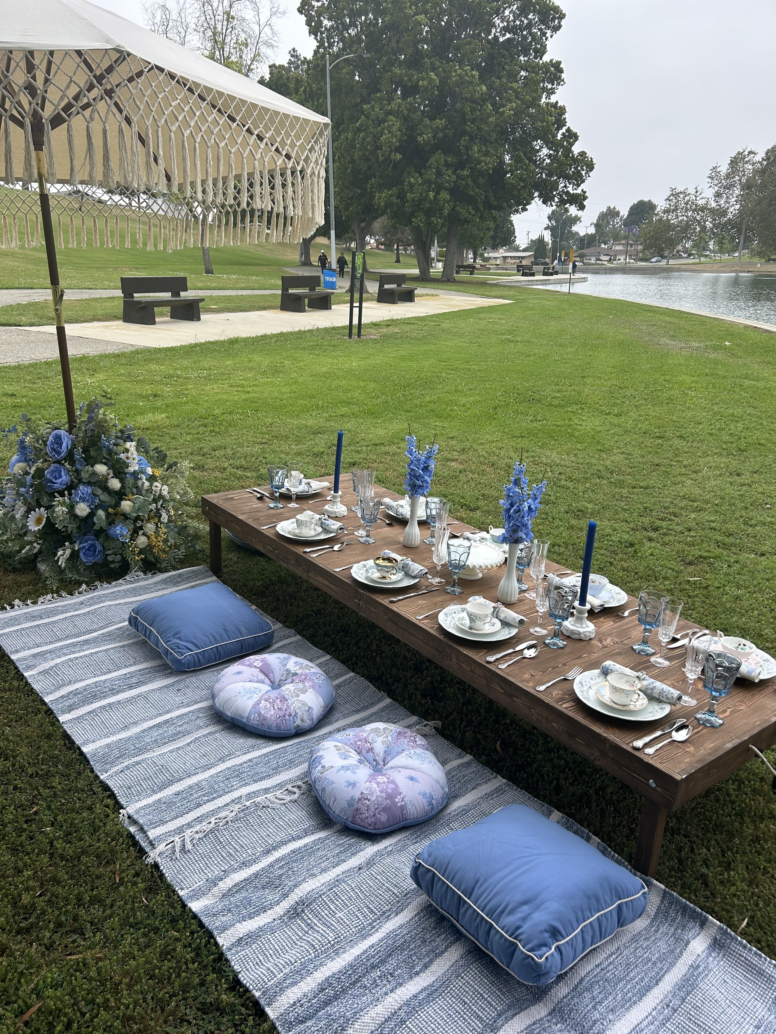 Outdoor picnic setup with a low wooden table on a grassy area, covered with plates, glasses, and floral arrangements. Blue and white cushions are on a striped blanket for seating, and a tent with fringe provides shade. A park setting with pathways, benches, and a body of water is visible in the background.