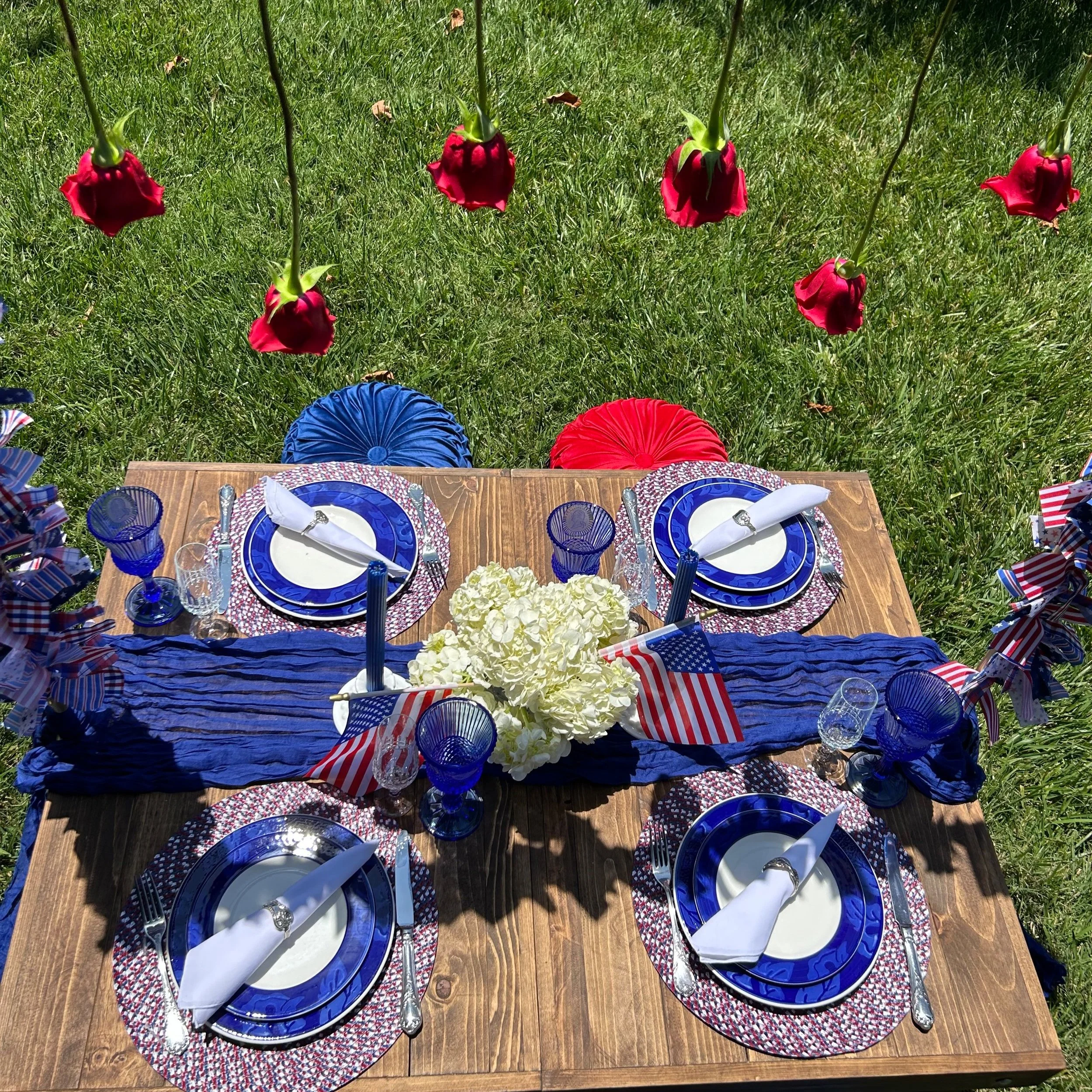 A patriotic-themed outdoor table setting on grass, featuring blue and white plates with red, white, and blue patterns. Red roses hang above. The table has blue glassware, white napkins, silver cutlery, and American flags. White hydrangeas and a blue fabric runner are centerpiece decorations. Red and blue seating cushions are visible.