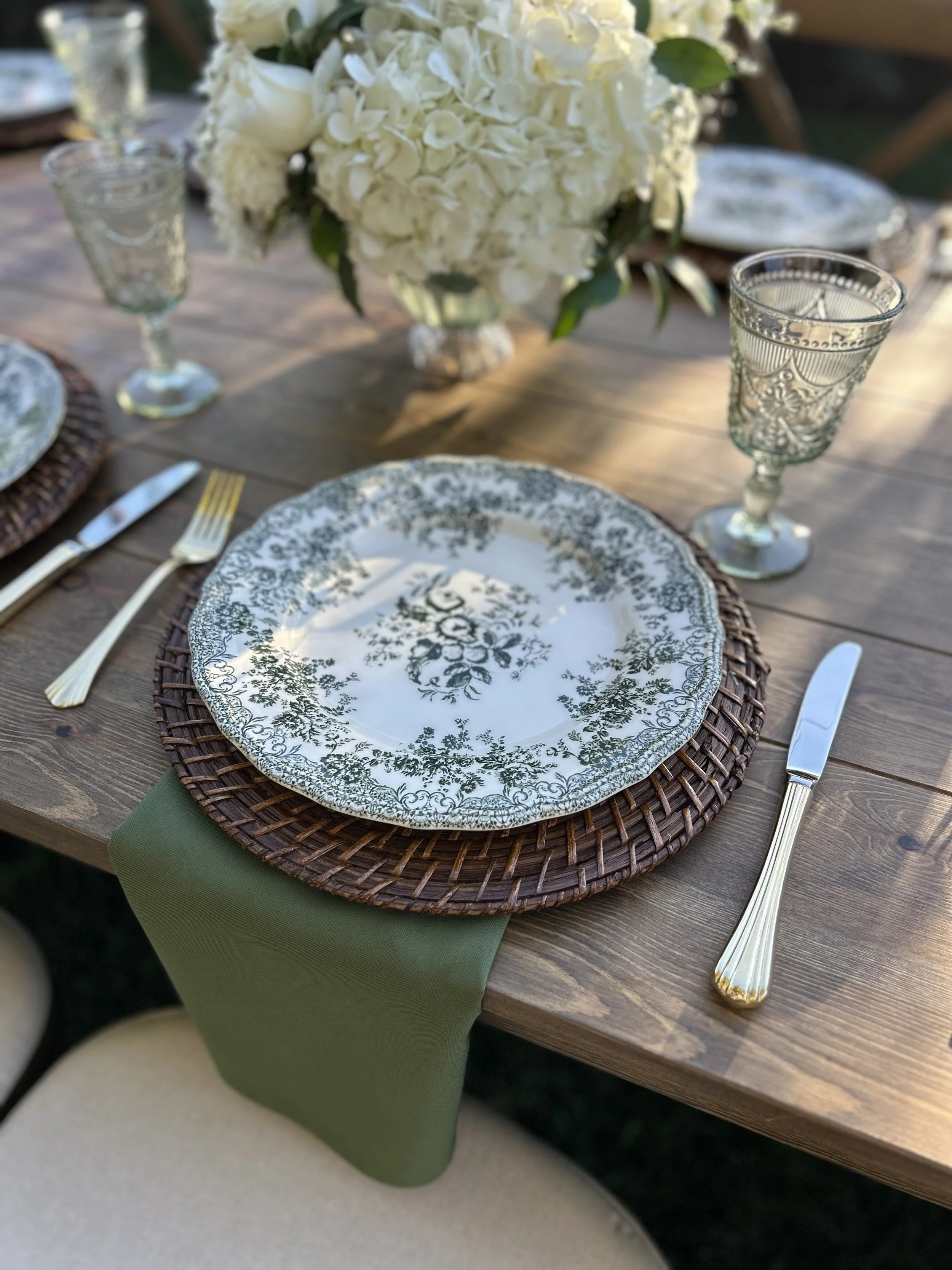 A table setting with a floral plate, gold fork and knife, green napkin, and a large bouquet of white flowers in a vase.