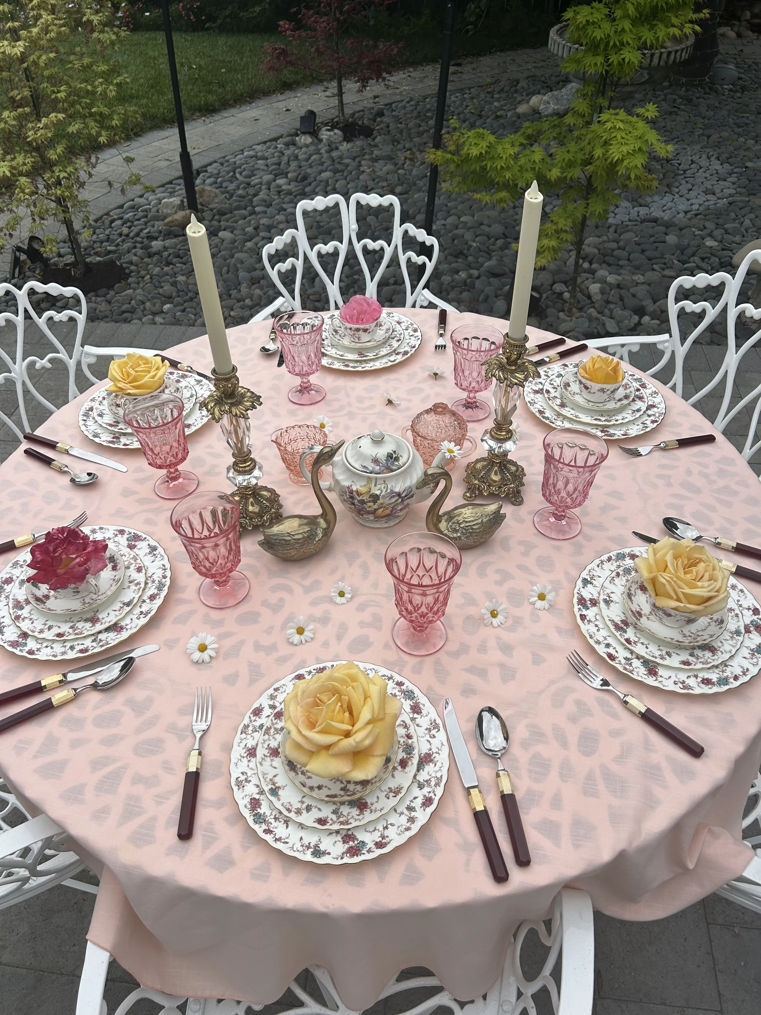 Elegant outdoor table setting with vintage floral china, pink glassware, and rose decorations. Features ornate candlesticks and decorative swans on a pink tablecloth. Surrounded by white chairs and garden scenery.