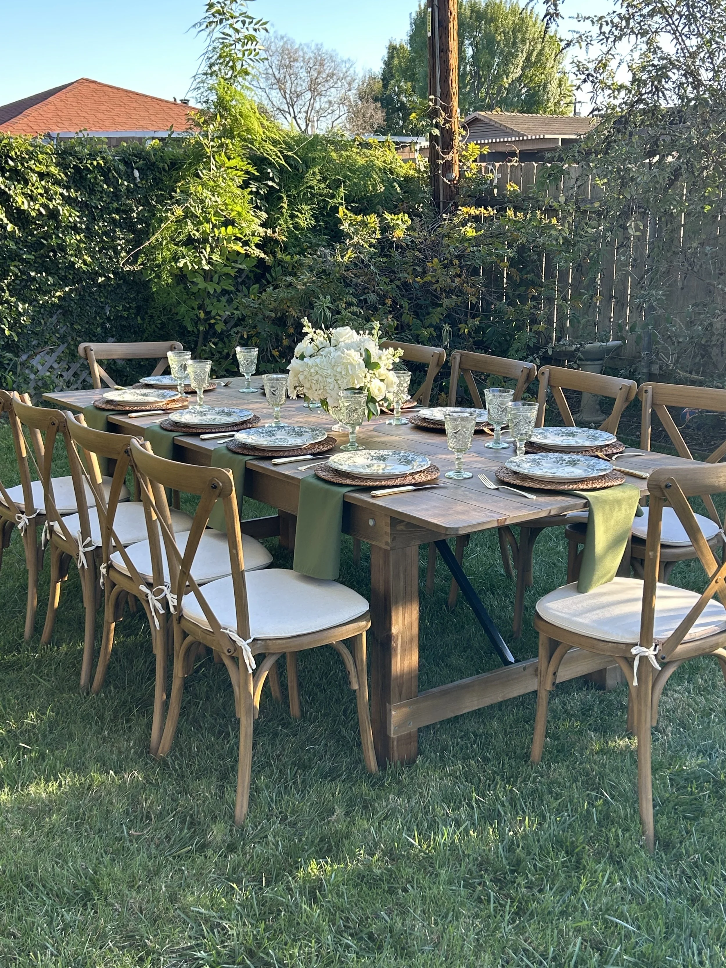 Outdoor dining table set for a gathering with plates, glasses, and a flower centerpiece, surrounded by wooden chairs on a grassy backyard with trees and a wooden fence.