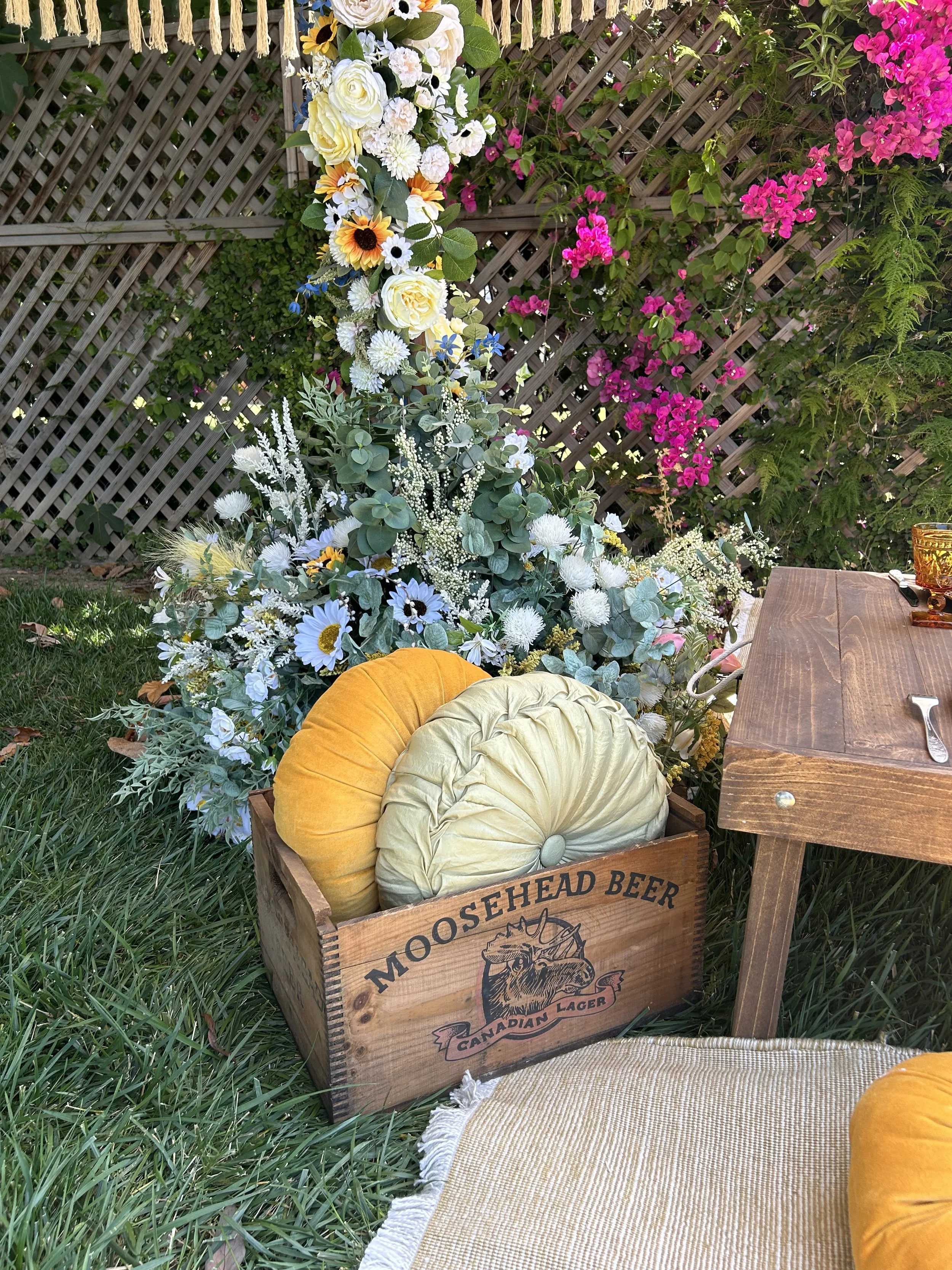 Outdoor setting with decorative floral arrangement and round cushions in a wooden Moosehead Beer crate. Vibrant pink, yellow, and white flowers with green foliage. Wooden table and wicker mat in the foreground.