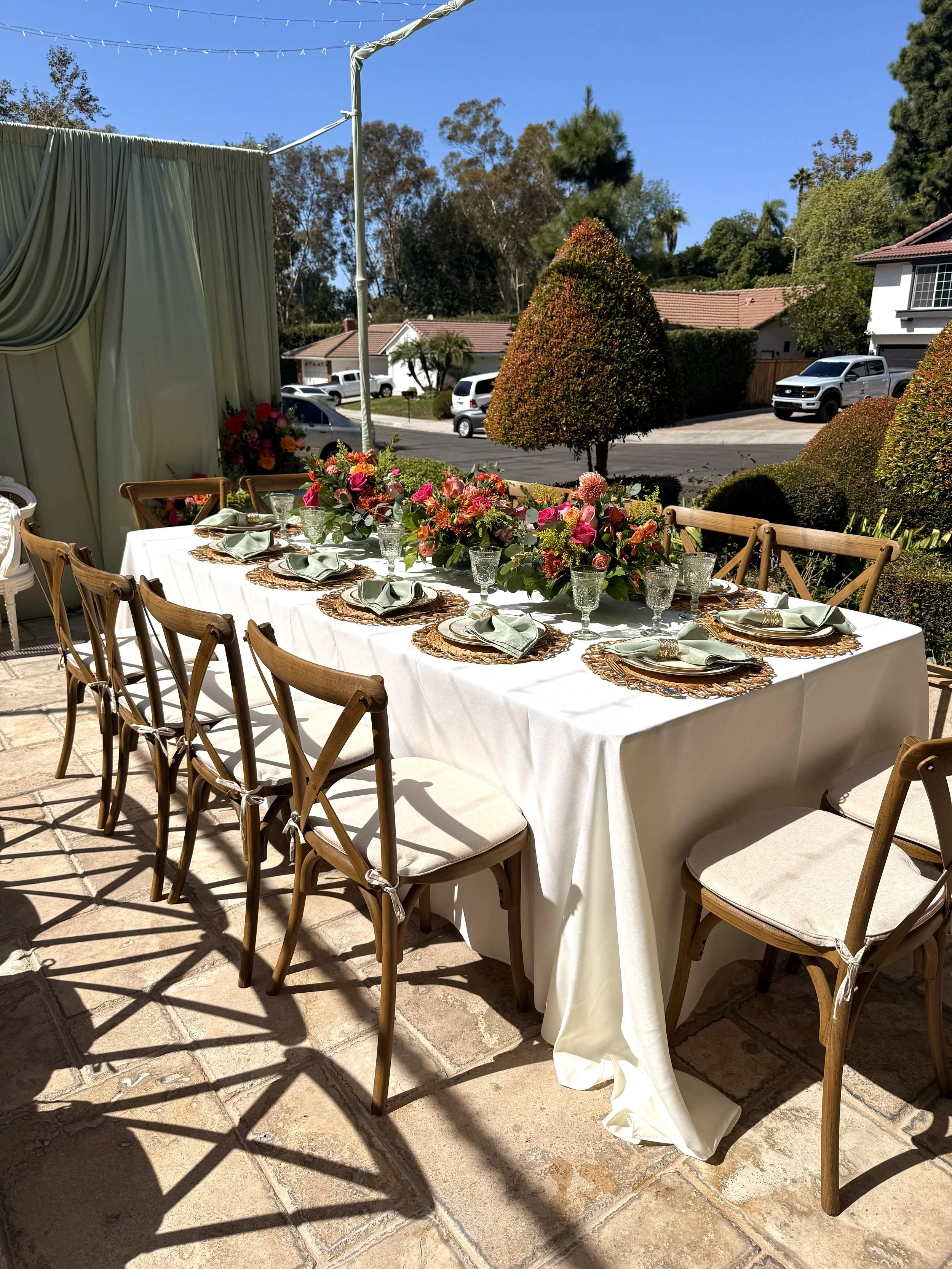 A long rectangular outdoor dining table set for a meal, with a white tablecloth, floral centerpieces, and place settings with china, silverware, and napkins. The table is surrounded by wooden chairs with cushions, and the setting is under a tent on a patio with a view of a residential neighborhood with trees and parked cars.
