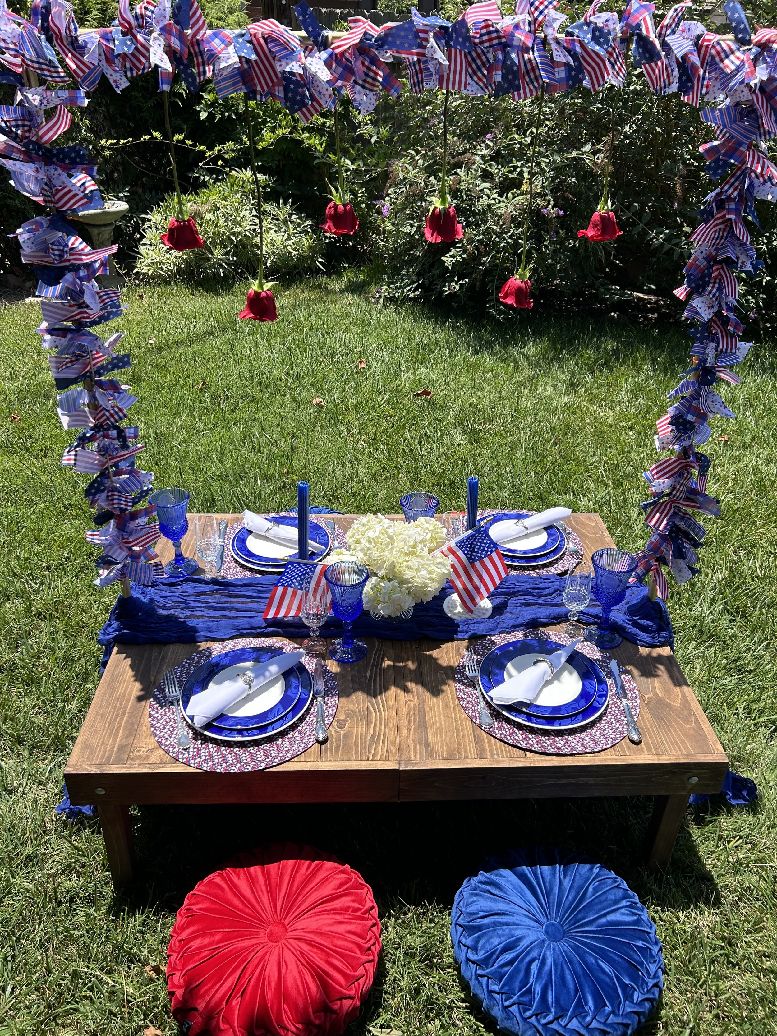 Patriotic outdoor table setting with American flags, blue and red décor, and hanging roses.