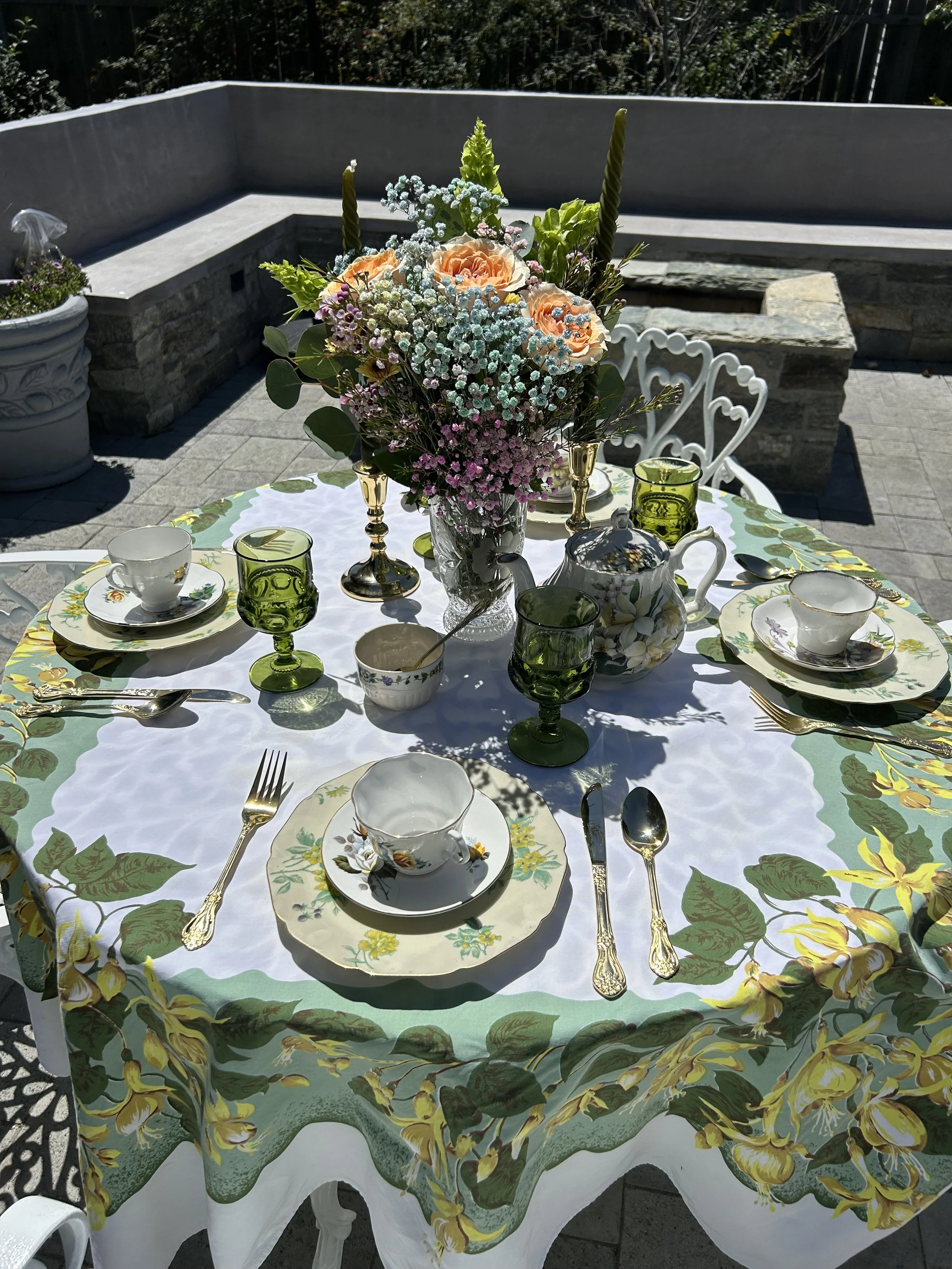 Outdoor table setting with floral tablecloth, tea set, green glassware, and a bouquet of flowers.