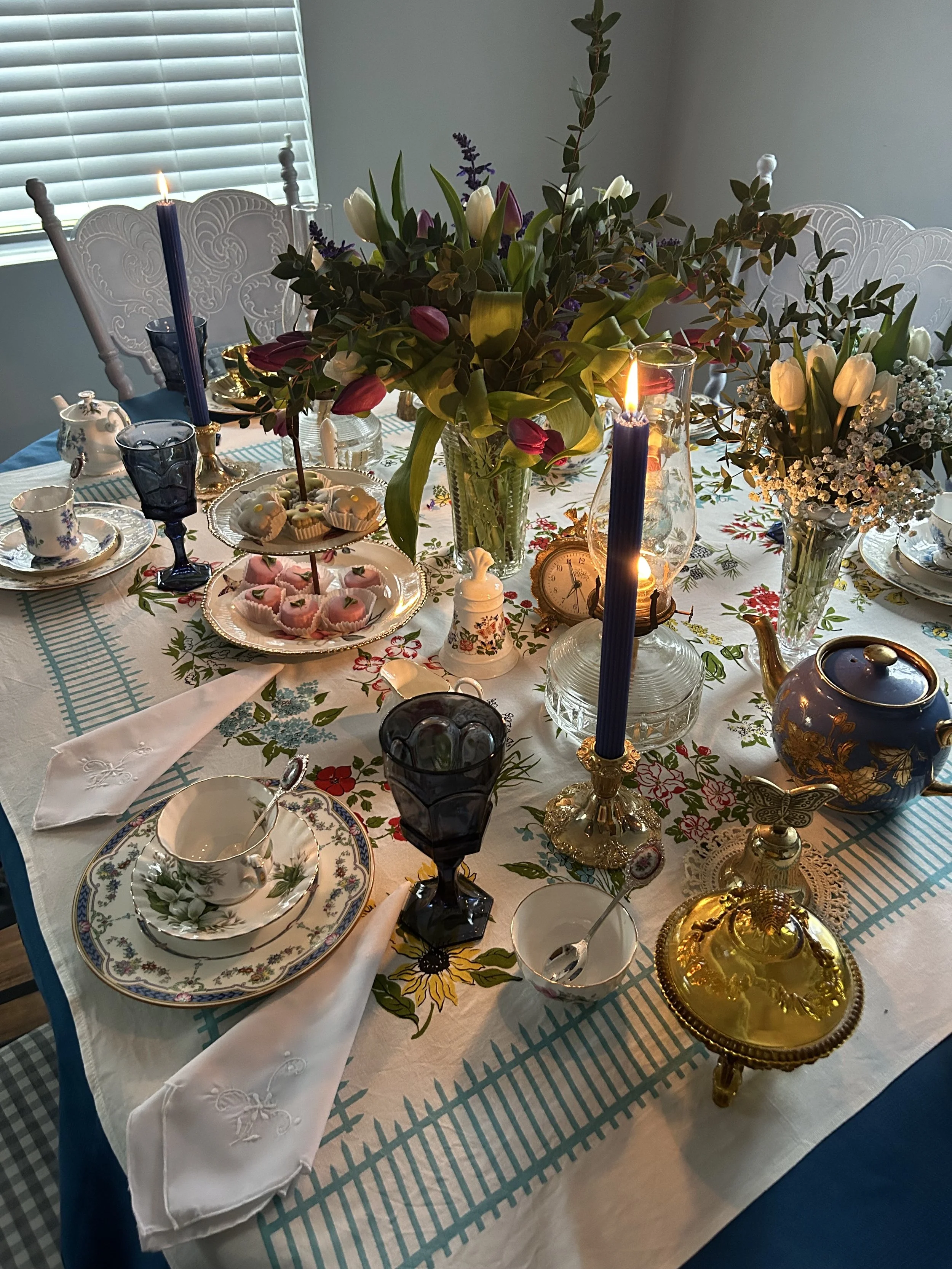 Elegant table setting with floral tablecloth, candlelit with blue candles, featuring vintage tea cups, plates, blue goblets, floral centerpieces with tulips, and an assortment of pastries and tea accessories.