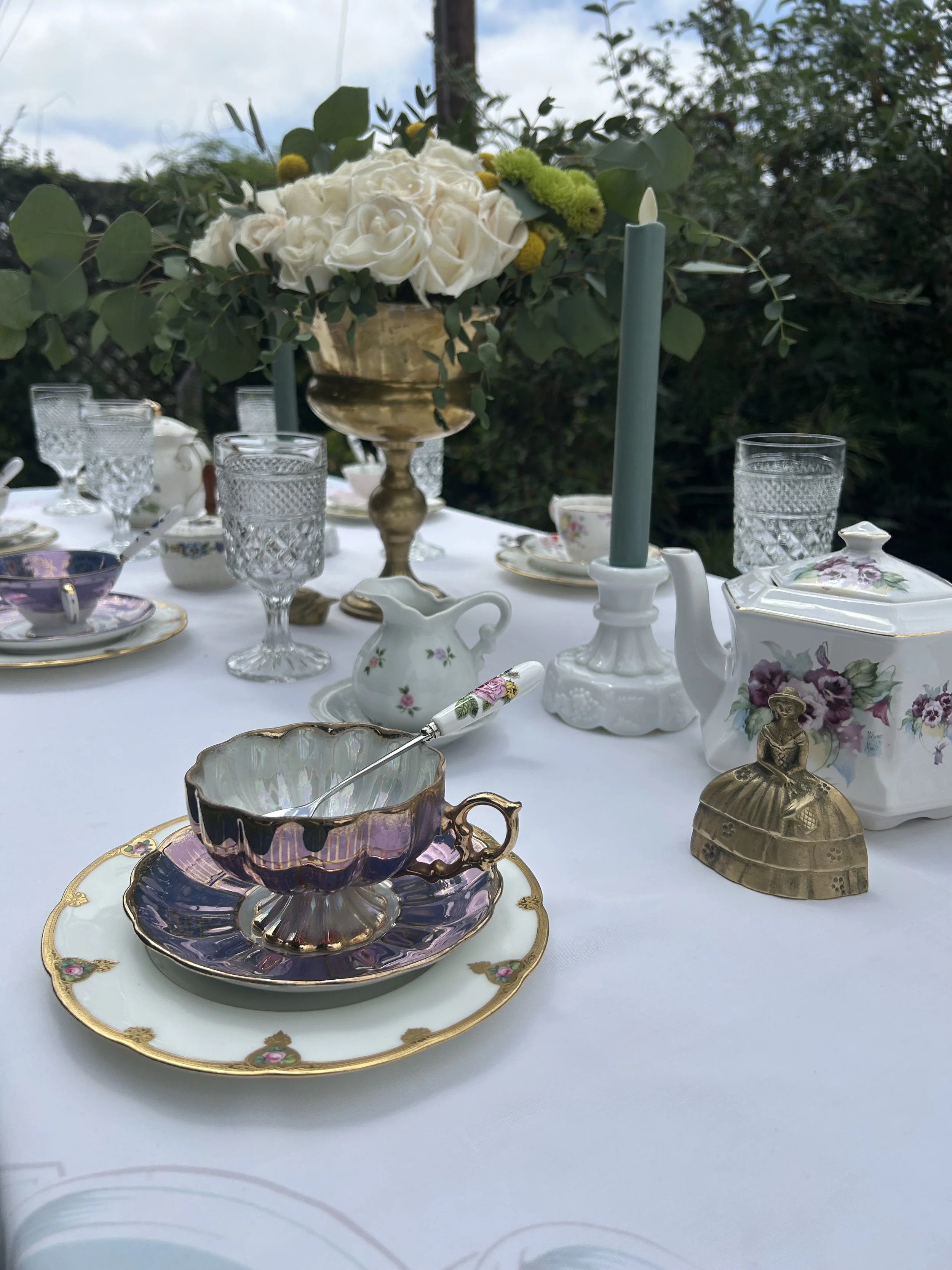Elegant outdoor tea party setup with vintage teacups, saucers, crystal glassware, and floral centerpiece on a white tablecloth.