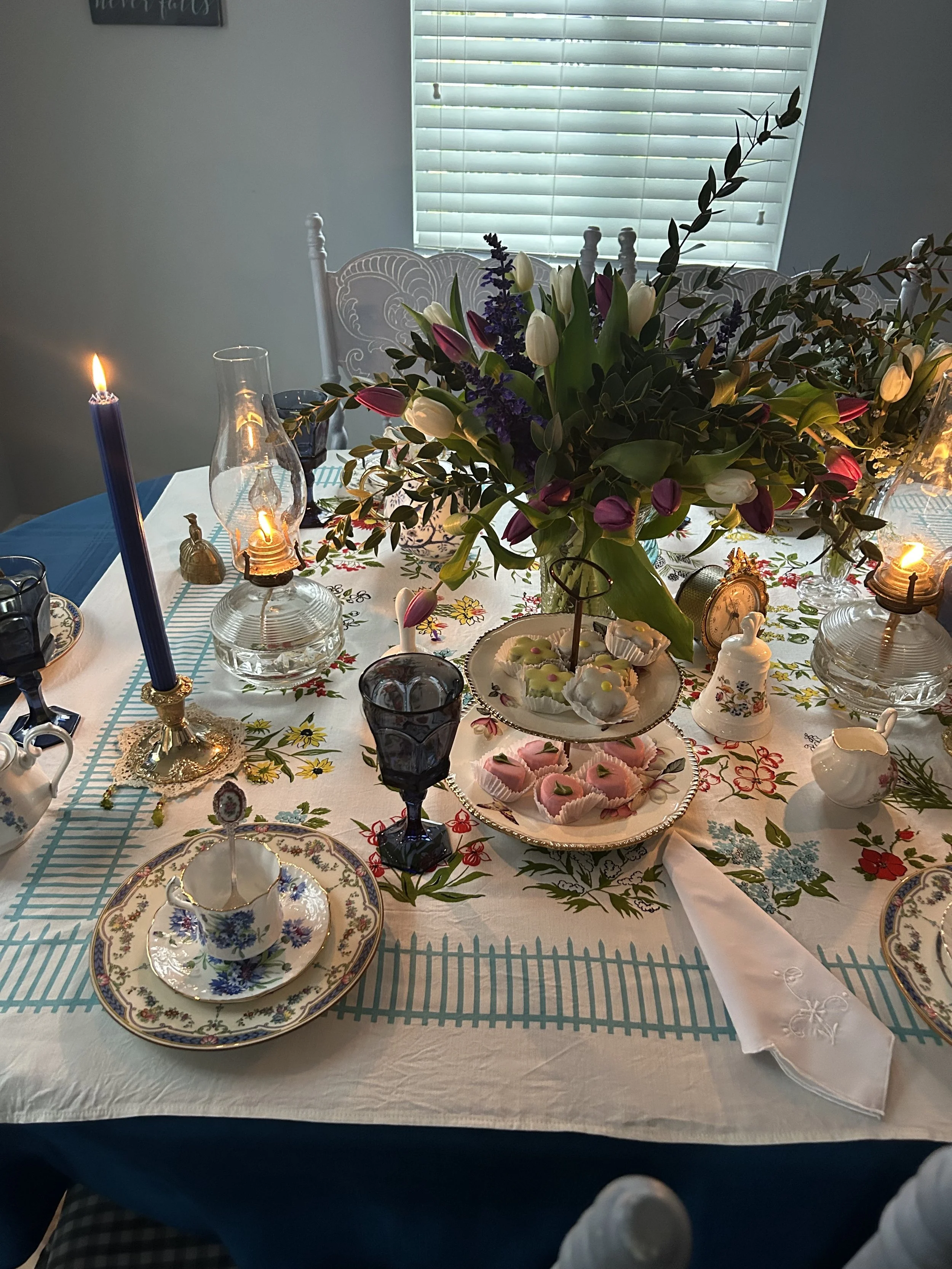 A dining table set for tea with floral-patterned china, a three-tiered cake stand with pastries, a large bouquet of purple and white flowers, and ornamental candles in holders.