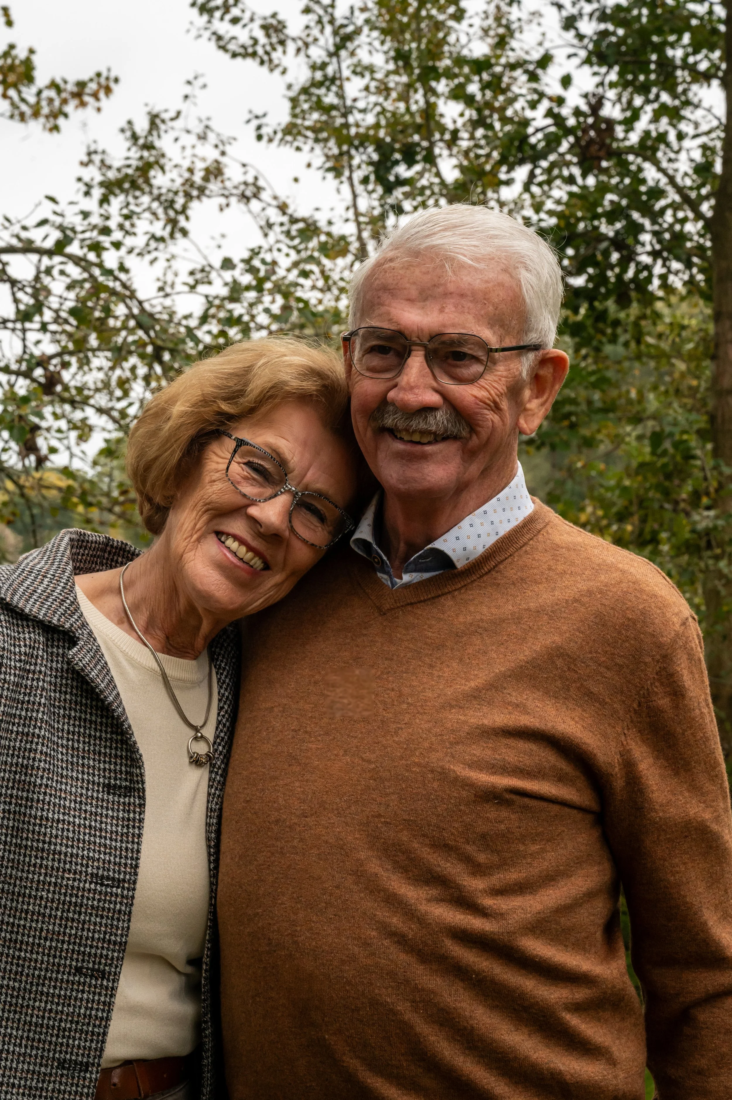 Een oudere man en vrouw, lachend en knuffelend, staan buiten met een achtergrond van bomen en een bewolkte hemel.