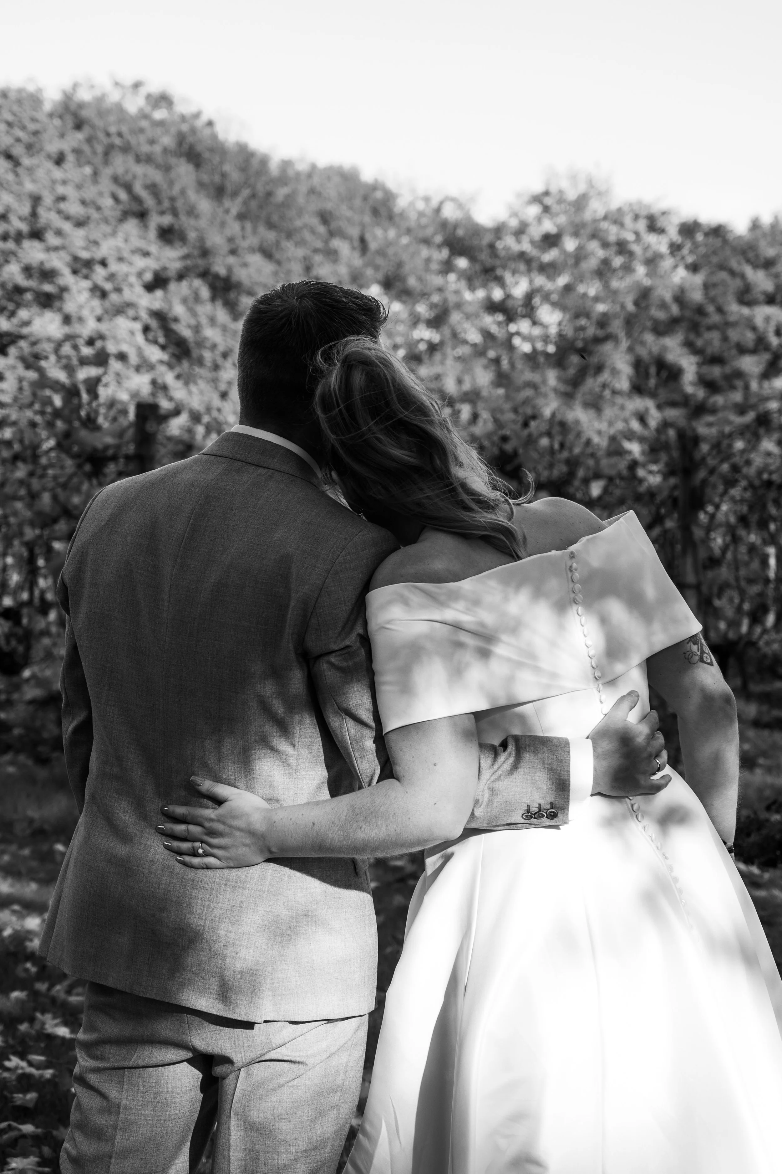 Zwart-wit foto van een stel dat elkaar omarmt op een natuurlijke achtergrond, mogelijk op een buitenbruiloft.