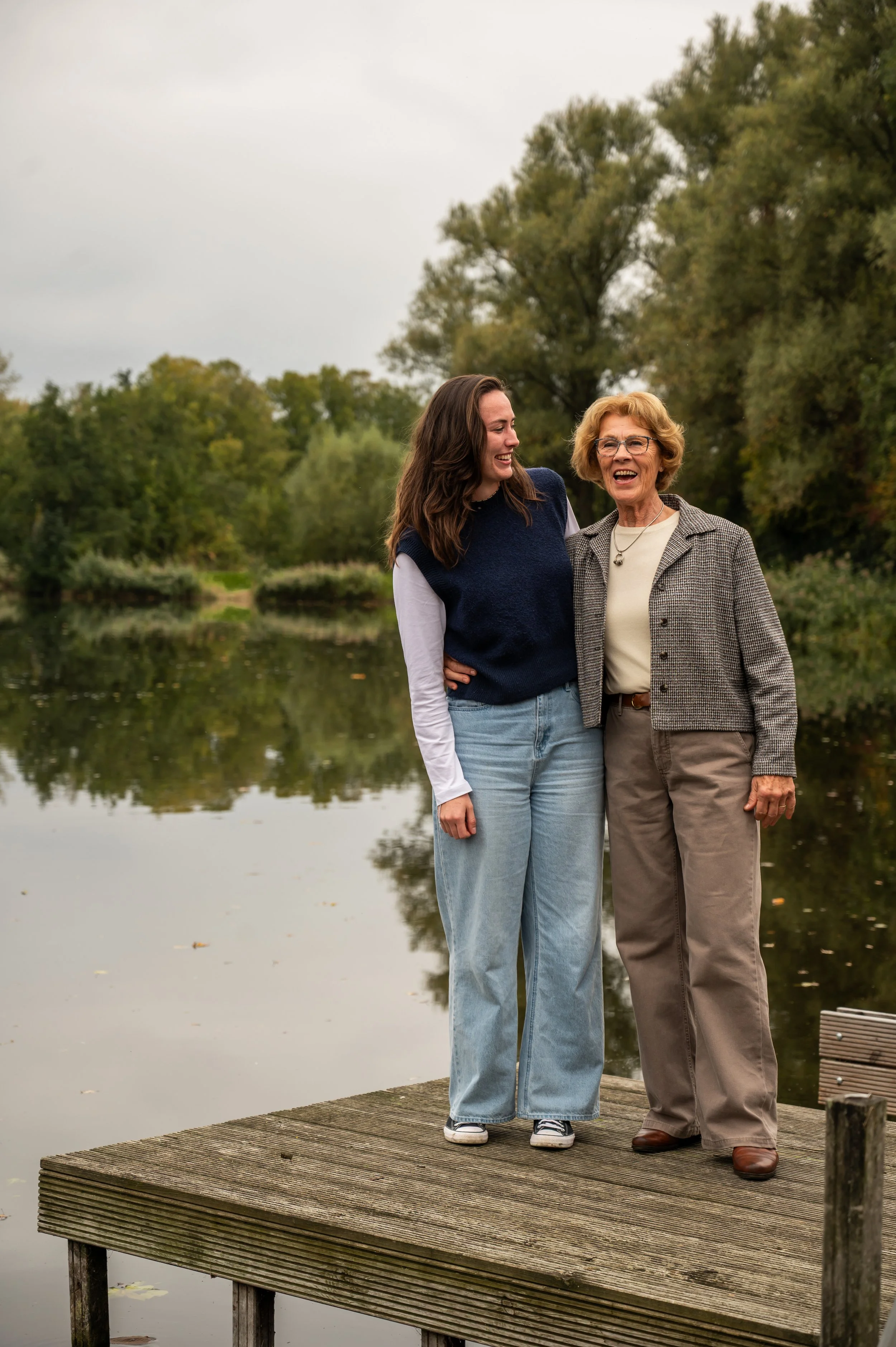 Twee vrouwen staan op een houten dok aan een meer, lachen en praten, met bomen en een bewolkte hemel op de achtergrond.