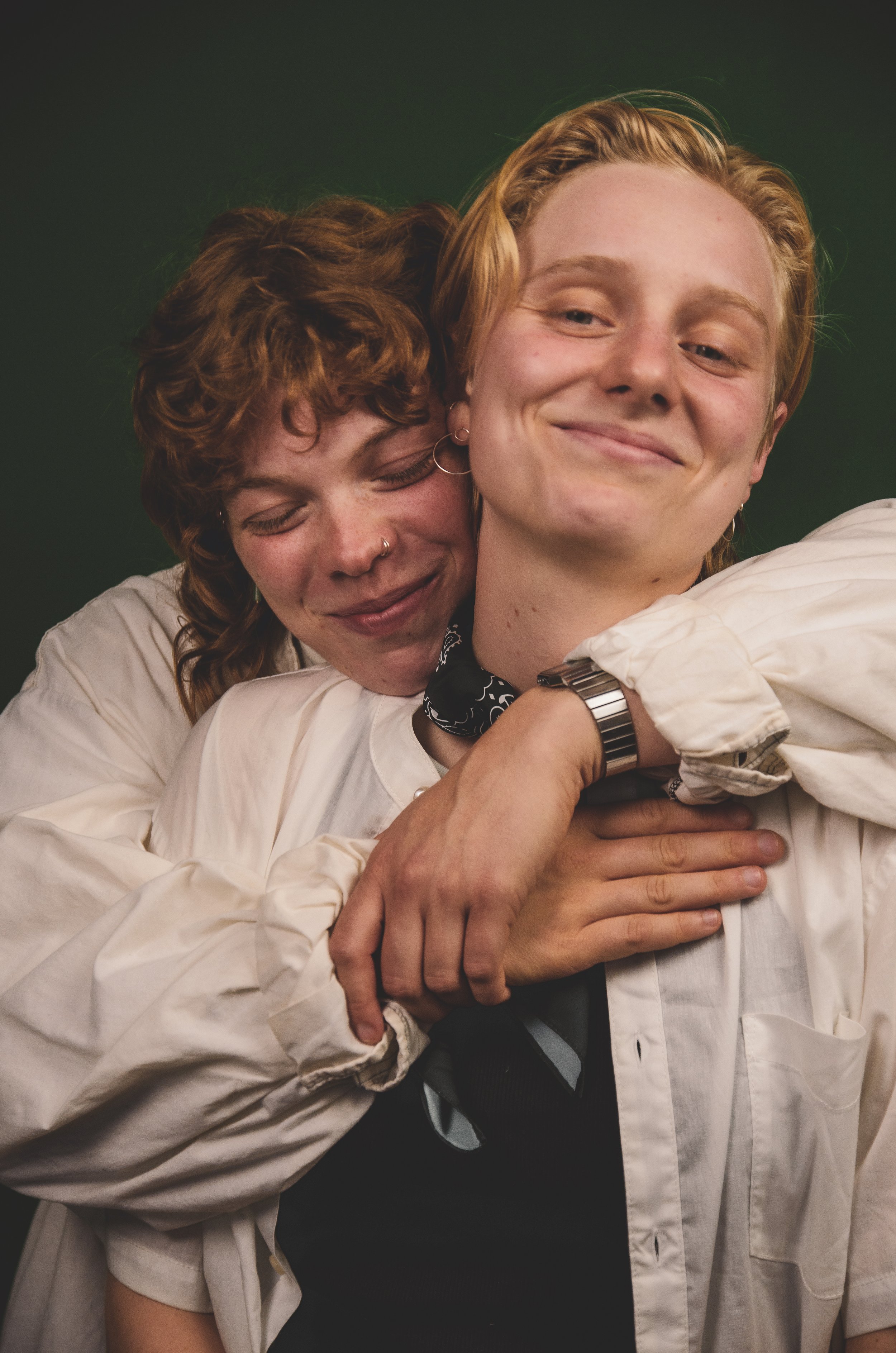 Two people embracing, one smiling and the other with eyes closed, both wearing white shirts against a dark green background.