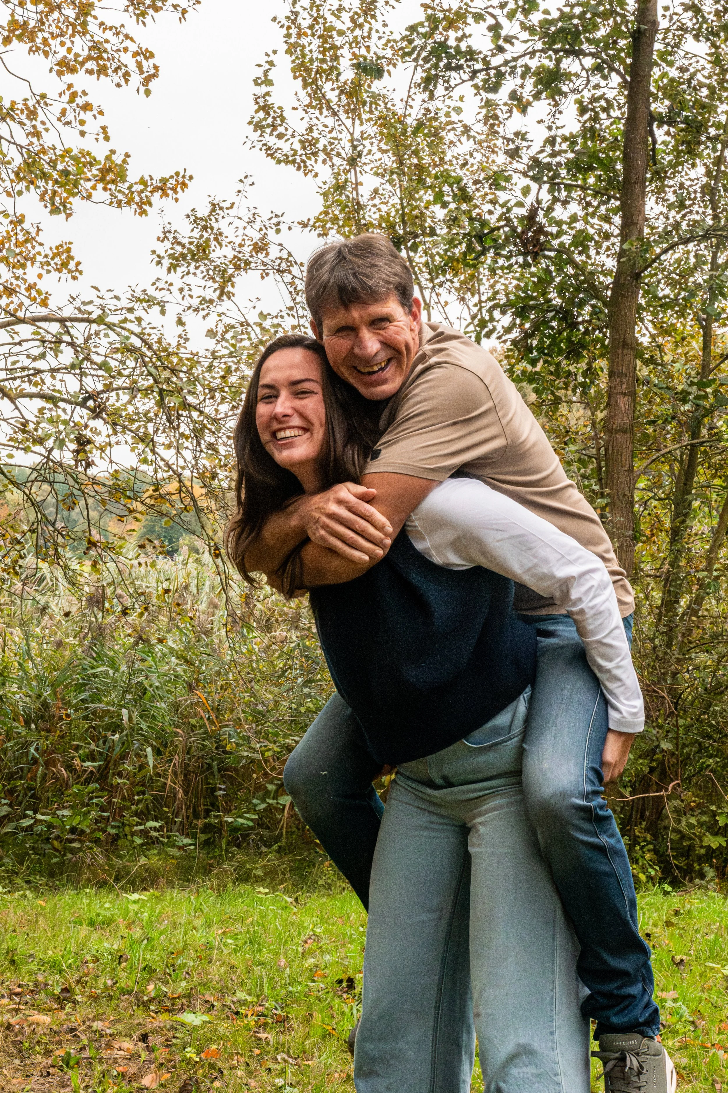 Twee mensen lachen en poseren samen in een bos, de man draagt de vrouw op zijn schouders.