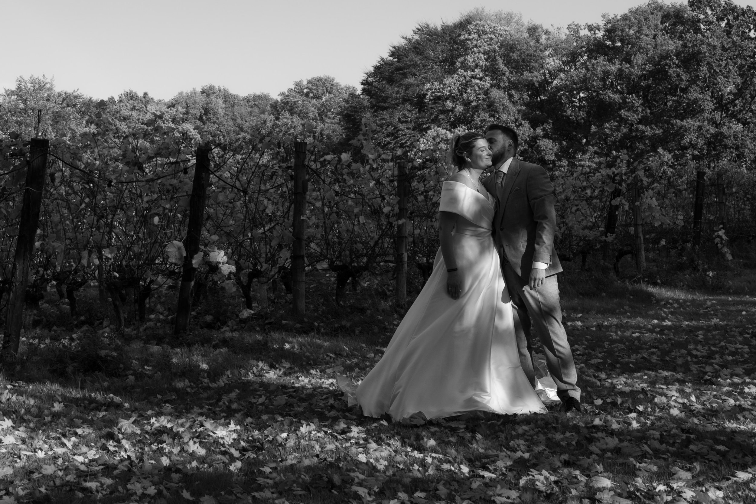 Zwart-wit foto van een bruid en bruidegom die elkaar kussen in een tuin met bomen en bladeren op de grond.