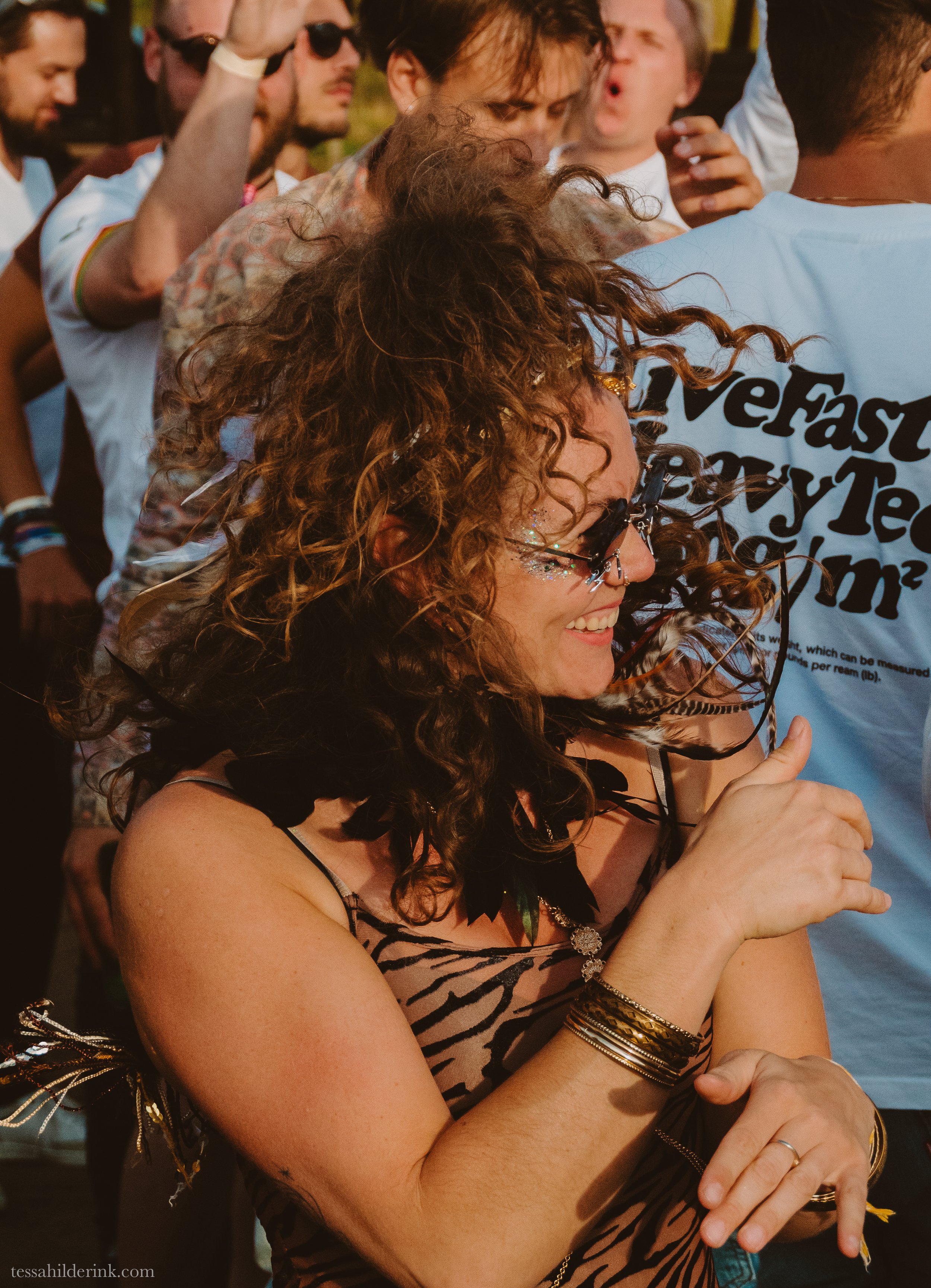 A lively outdoor dance party with a smiling woman wearing sunglasses and a zebra-print top, embellished with feathers and glitter.